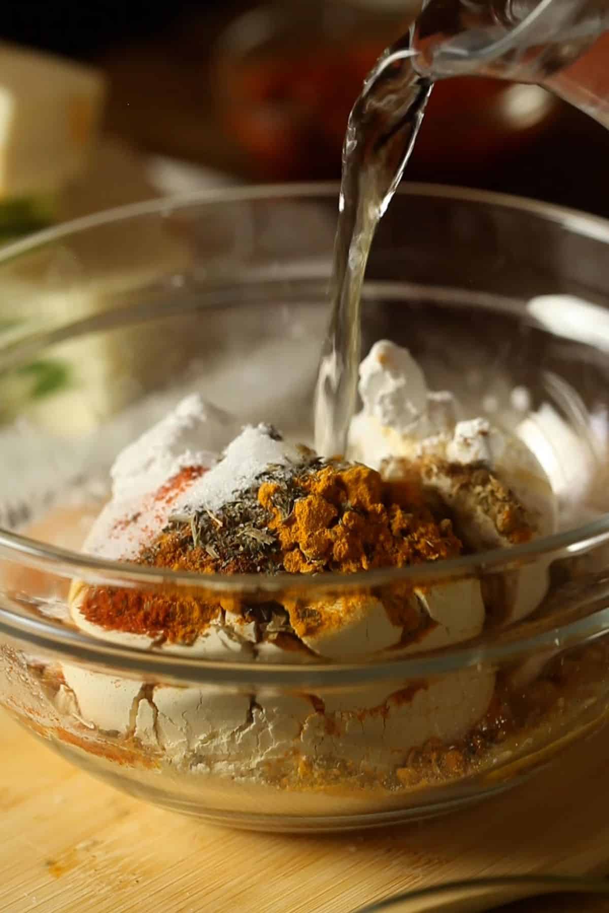 Ingredients for pakora batter are placed in a glass bowl and water is poured on them.