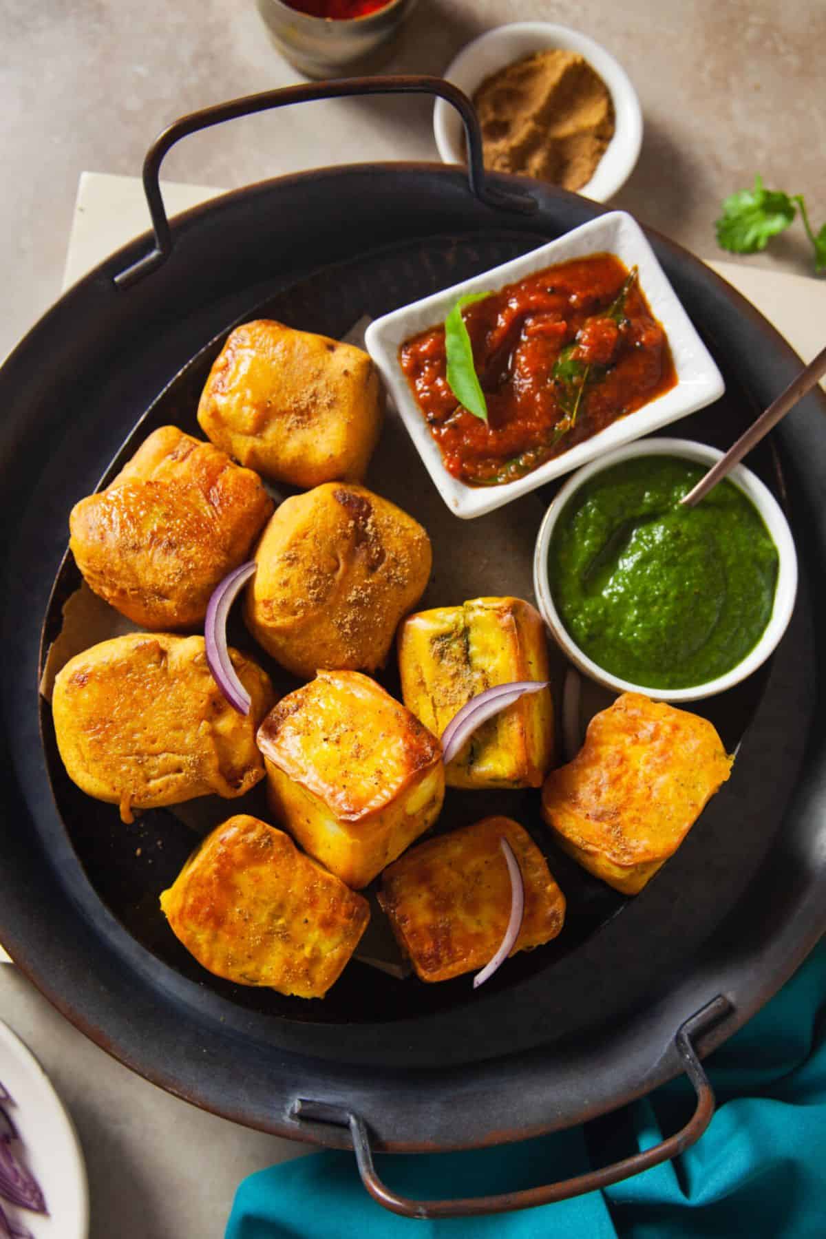 Paneer Pakoras placed on a black tray with bowls of green and tomato chutney.