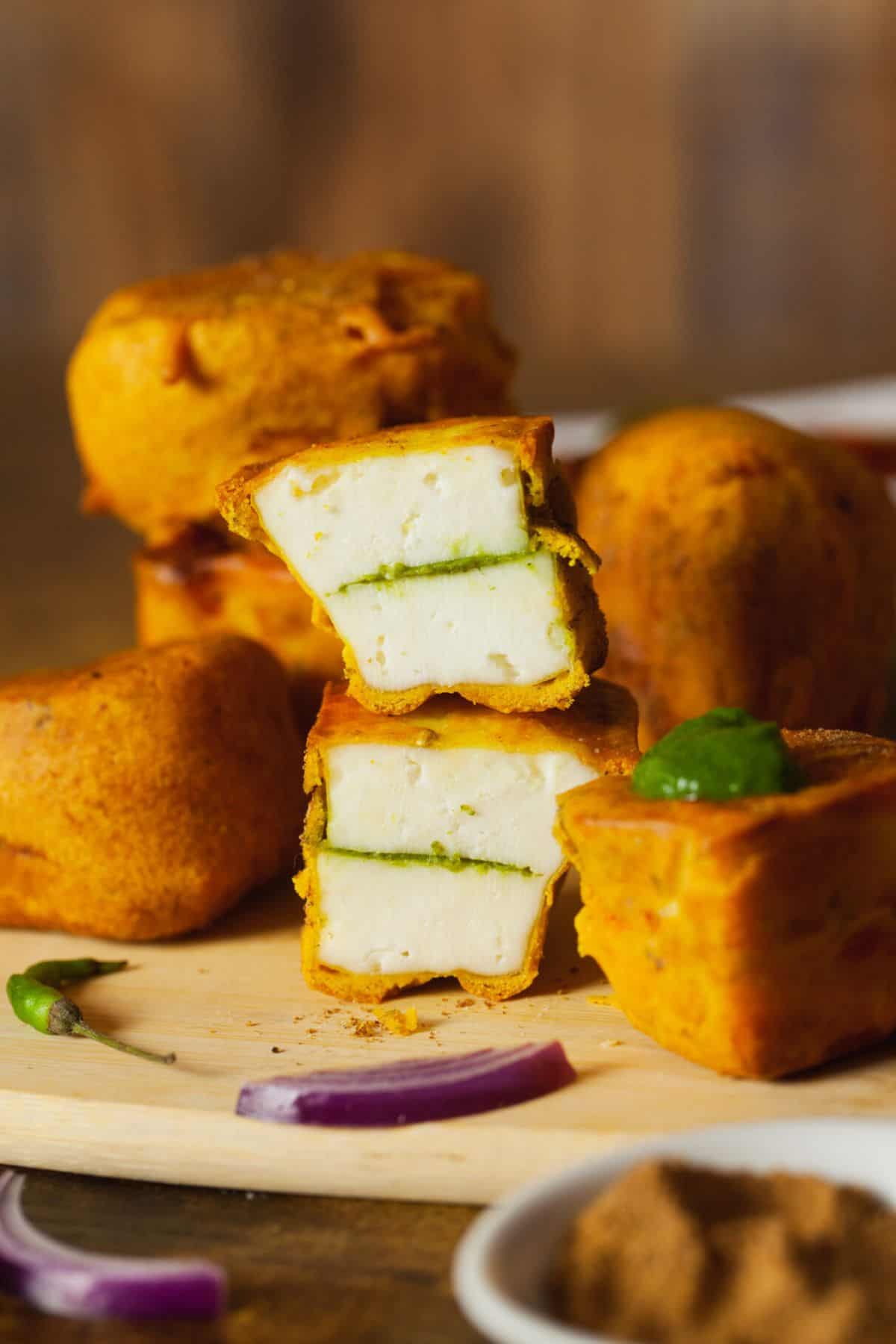 Cut up paneer pakora stacked on a wooden board to show chutney layer inside.