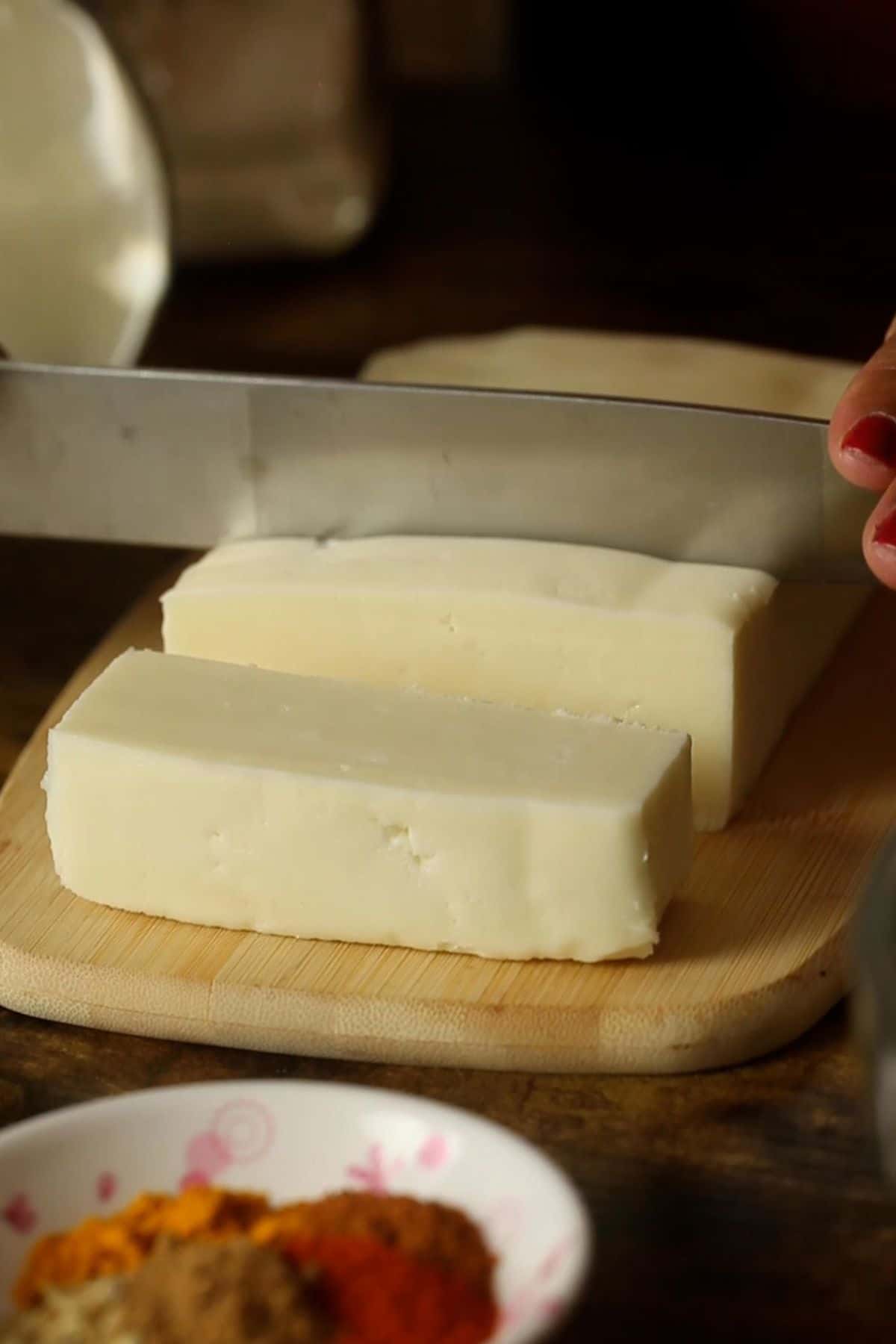 A block of paneer placed on a bamboo board and is being cut with a metal knife.