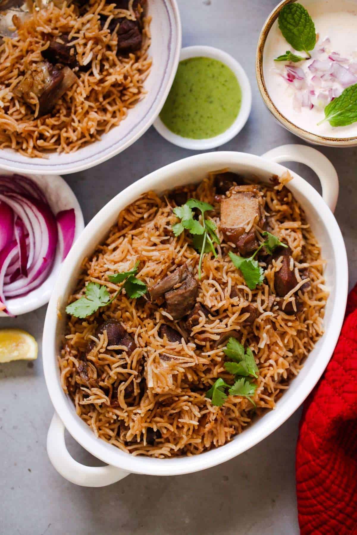 Gosht Pulao in an oval dish with onions, green chutney and raita yogurt.