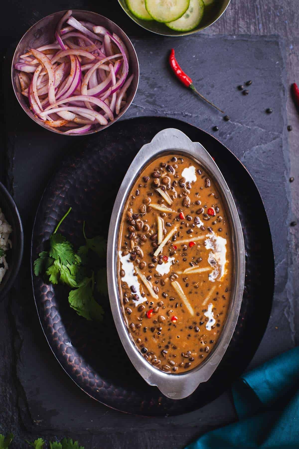 ITC Style Dal Bukhara in an oval dish with sliced onions, cucumbers and flatbreads. 