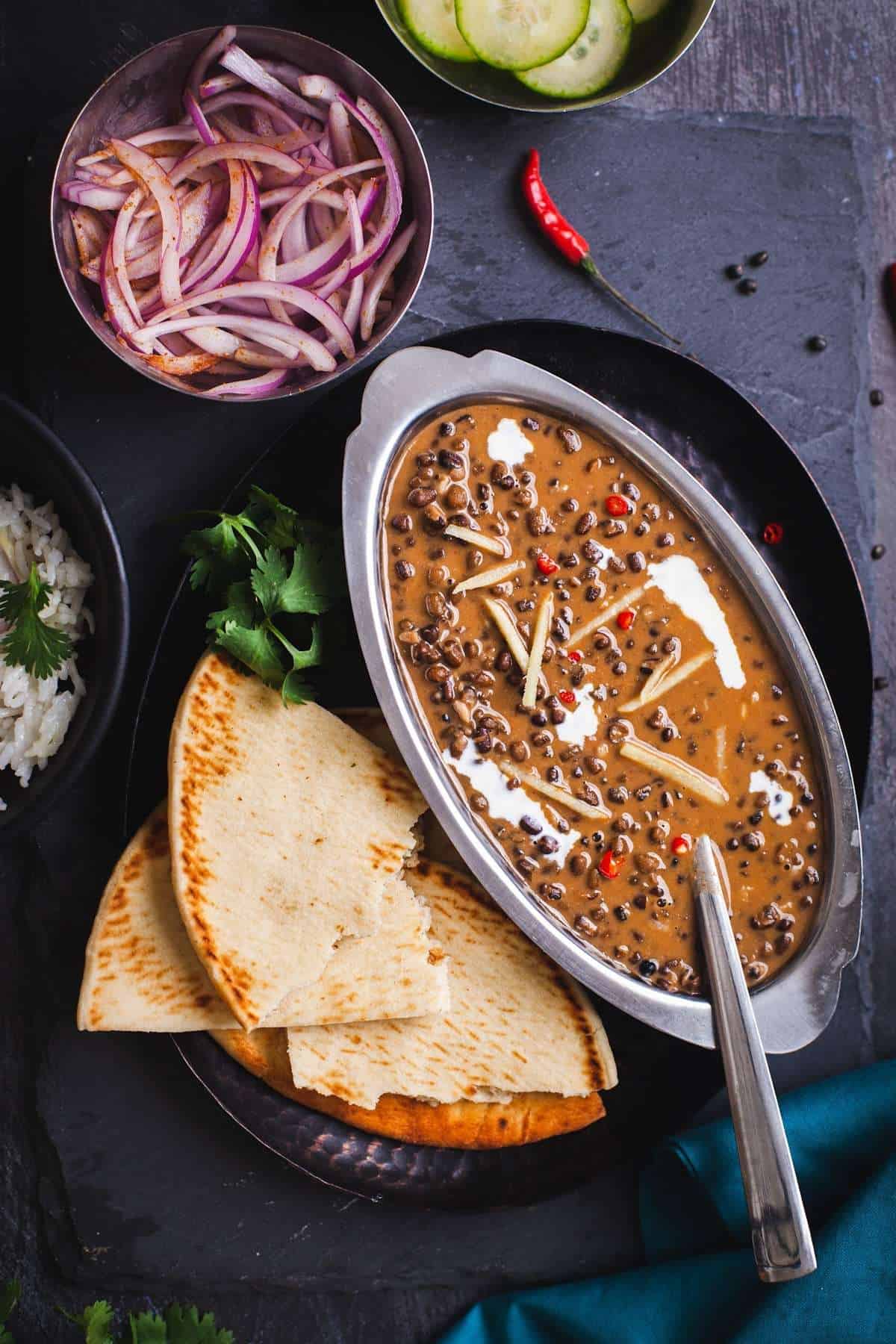 ITC Style Dal Bukhara in an oval dish with sliced onions, cucumbers and flatbreads.