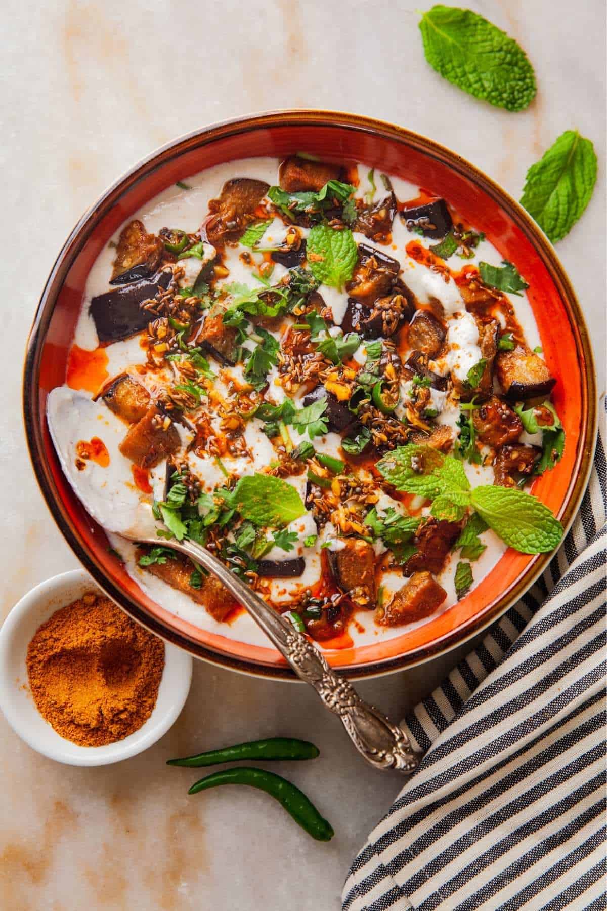 Dahi Baingan in a red shallow plate with toppings and tempering and sprinkle of chaat masala.