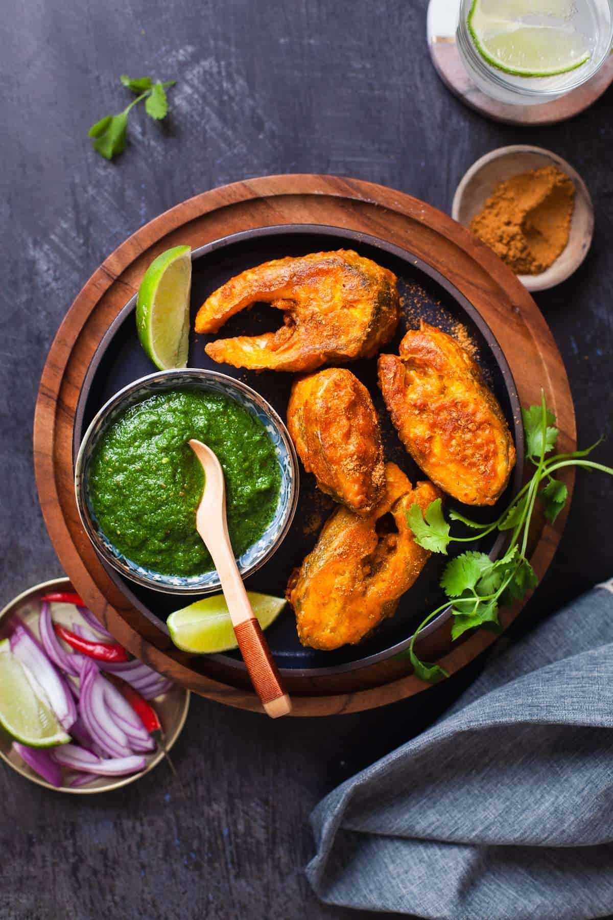 Amritsari Fish with fish steaks served with Green Chutney and sliced onions on a black plate.