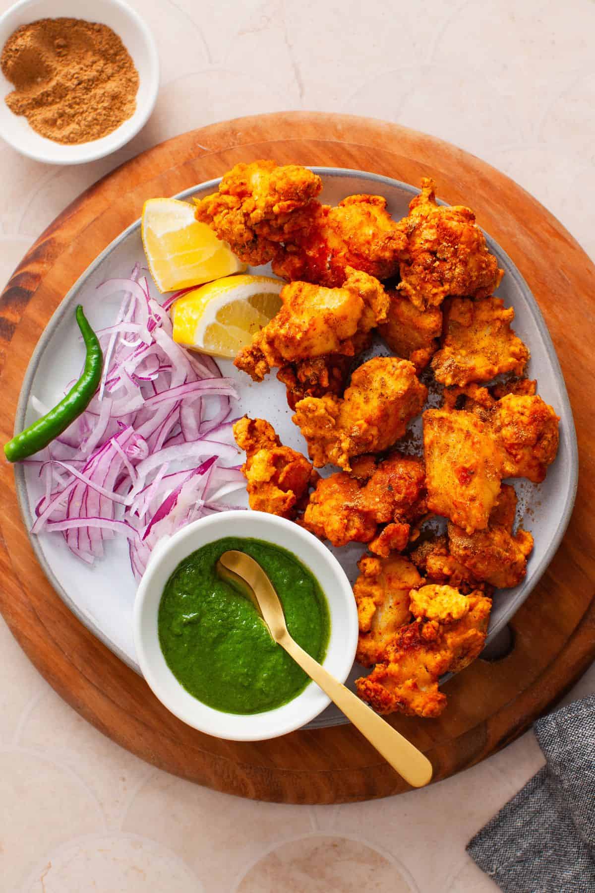 Fish pakora served on a white plate with green chutney,sliced onion and lemon.
