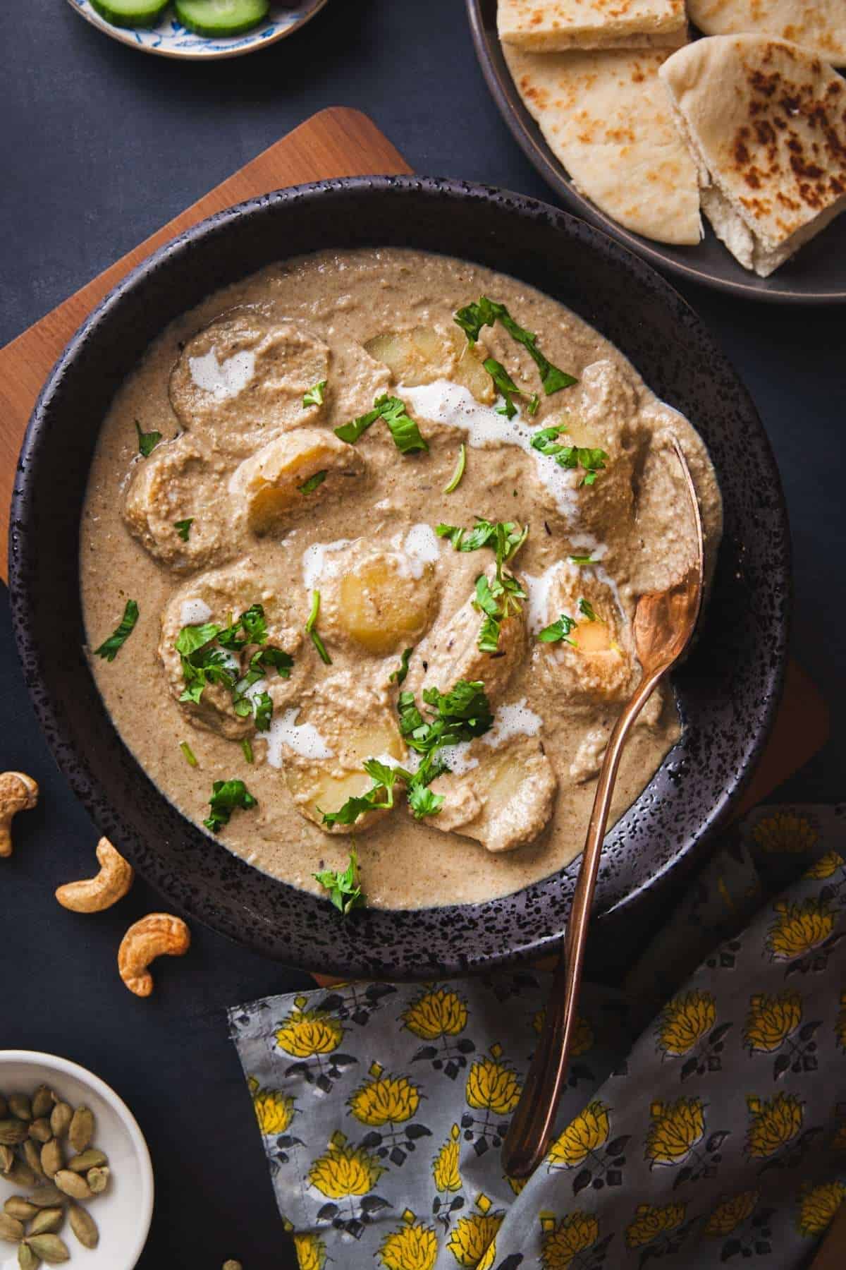 Malai Aloo in a black round dish served with naan and a salad of cucumber and onions.