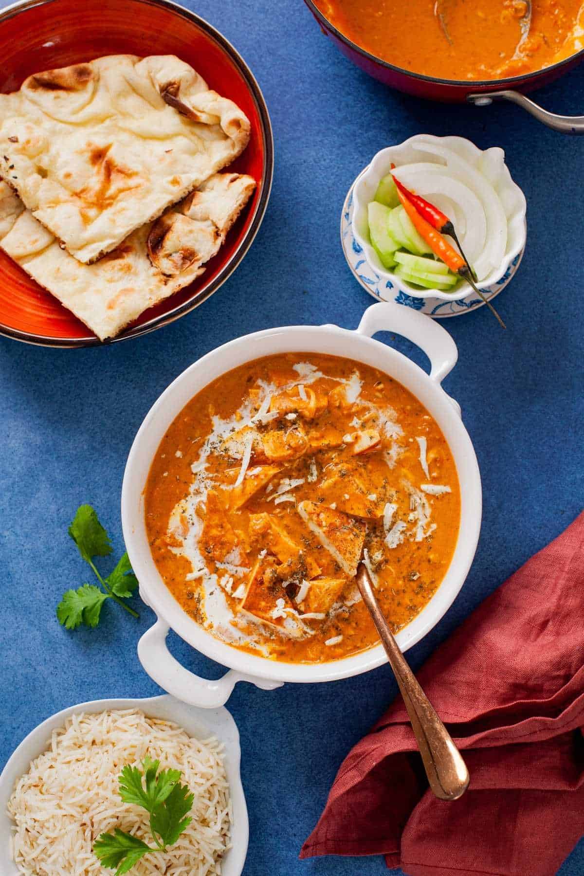 Paneer lababdar in a white dish placed on a denim blue background with cumin rice, naan and indian salad. 