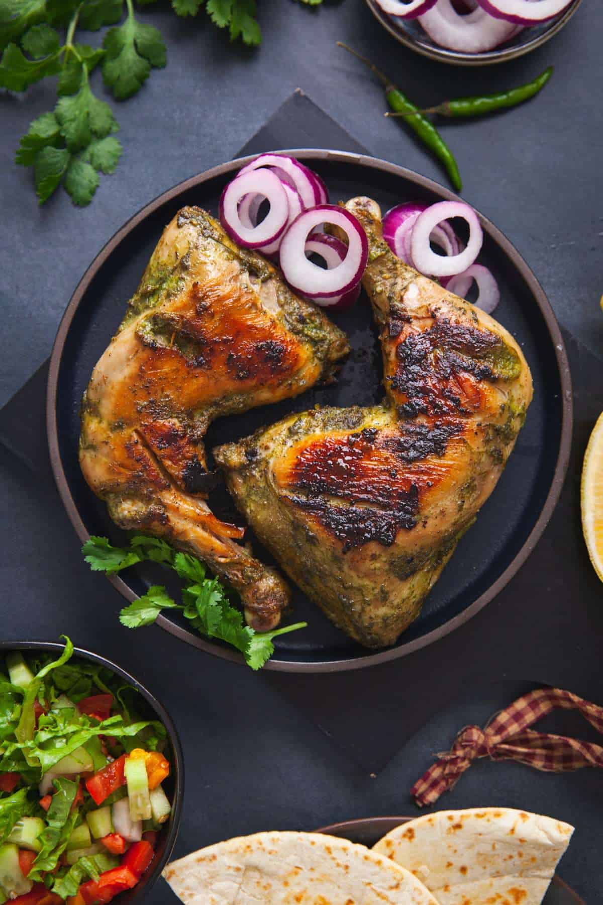 Hariyali Chicken leg quarters served on a black plate with onion rings, flatbreads and cucumber tomato salad. 