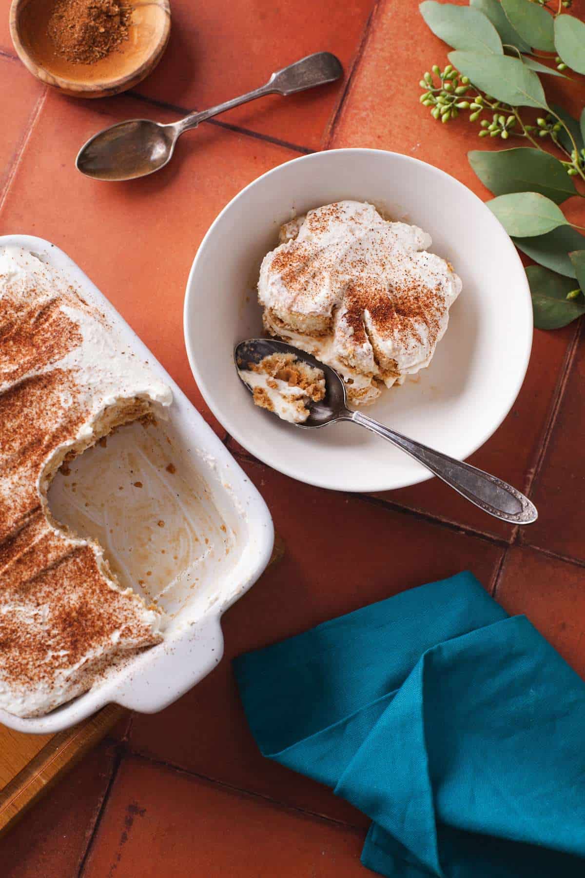 A slice of Chai Tiramisu in a white bowl and spices on a terra-cotta background.