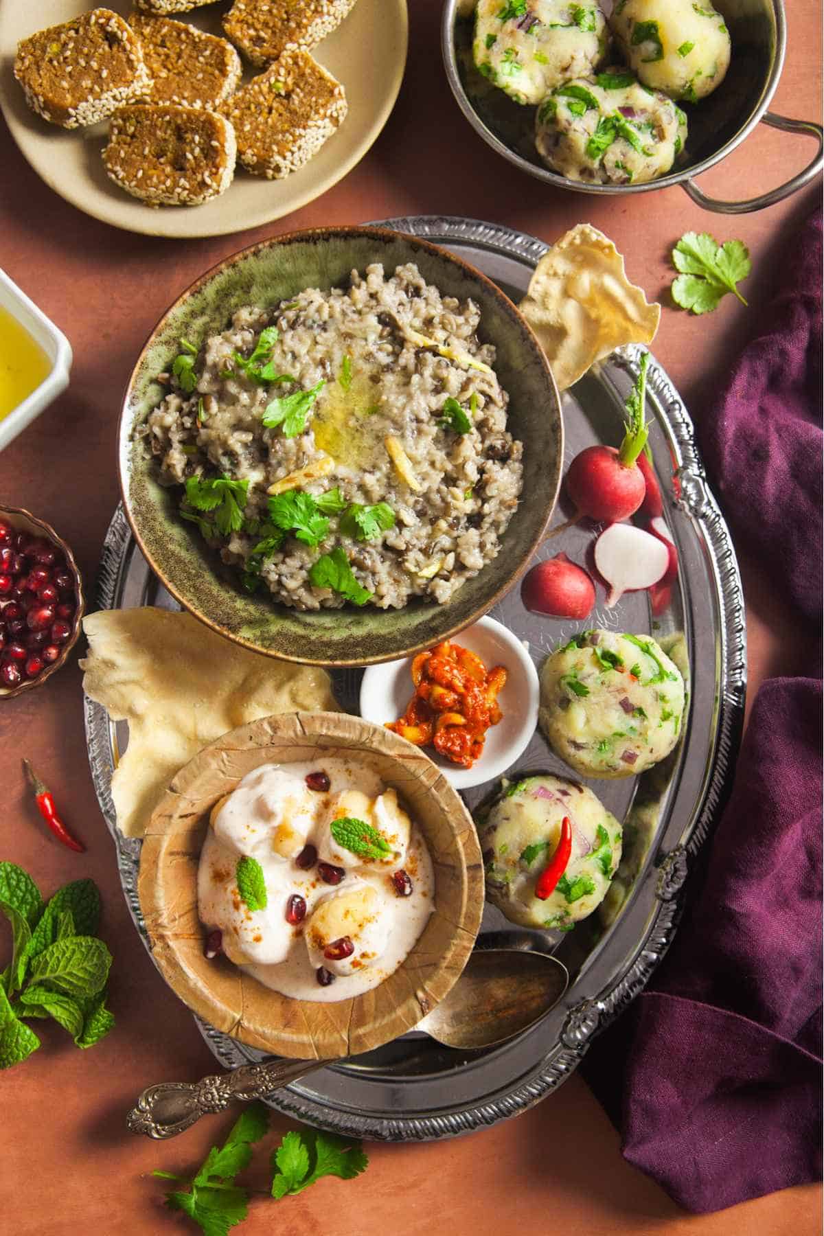 Makar Sankranti Khichdi Meal served on a oval steel plate with urad dal khichdi, aloo bharta, sesame laddus and dahi vada.