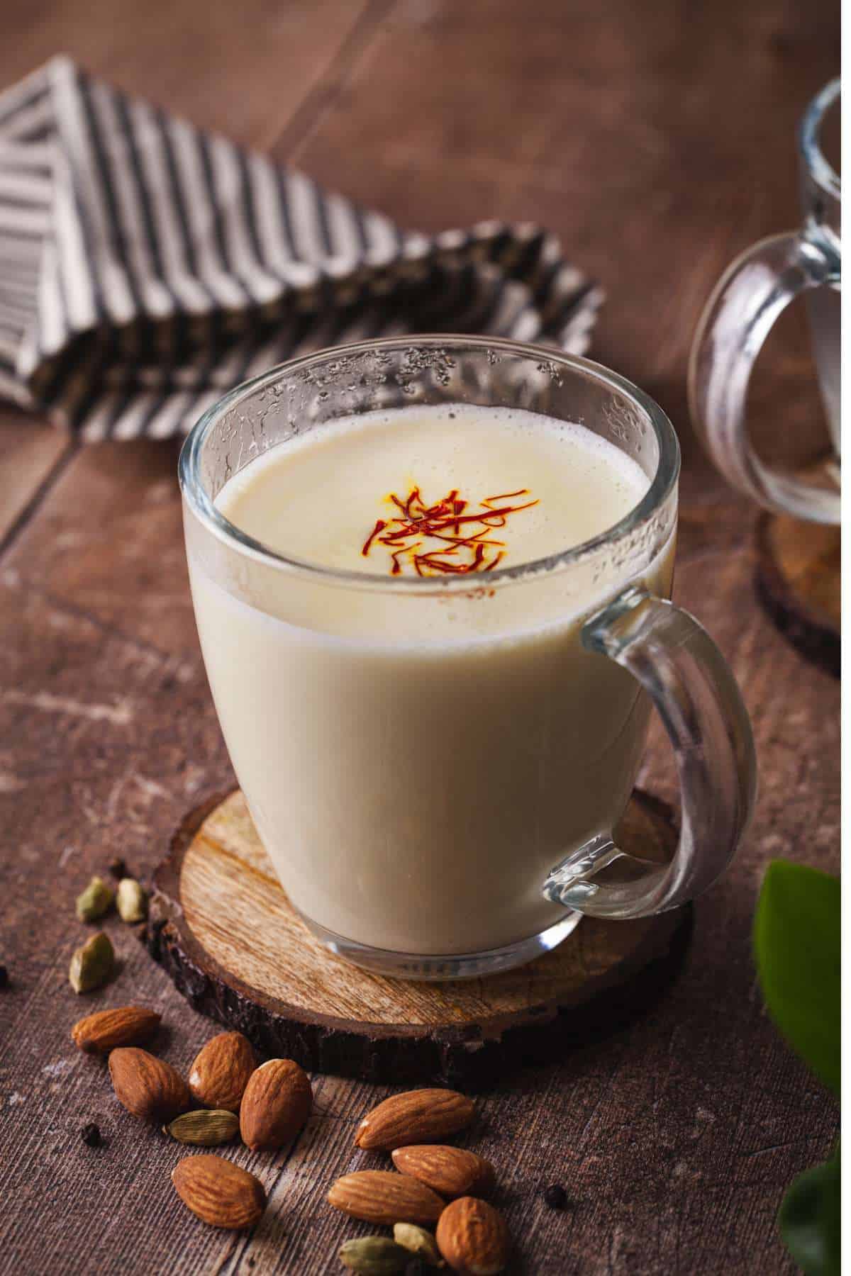 Badam Milk in a glass cup with almonds and cardamom scattered around.