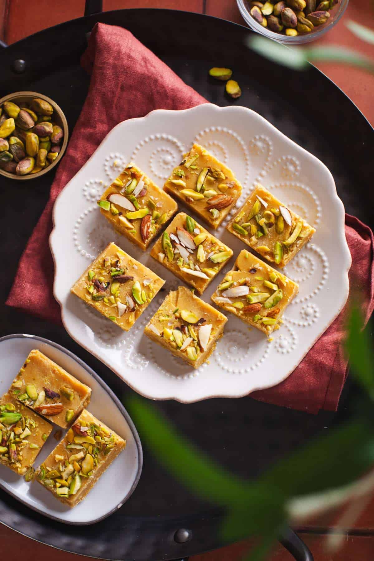Pieces of Besan Ki Barfi on a fluted white pale with a red cloth and small bowl of pistachios. 