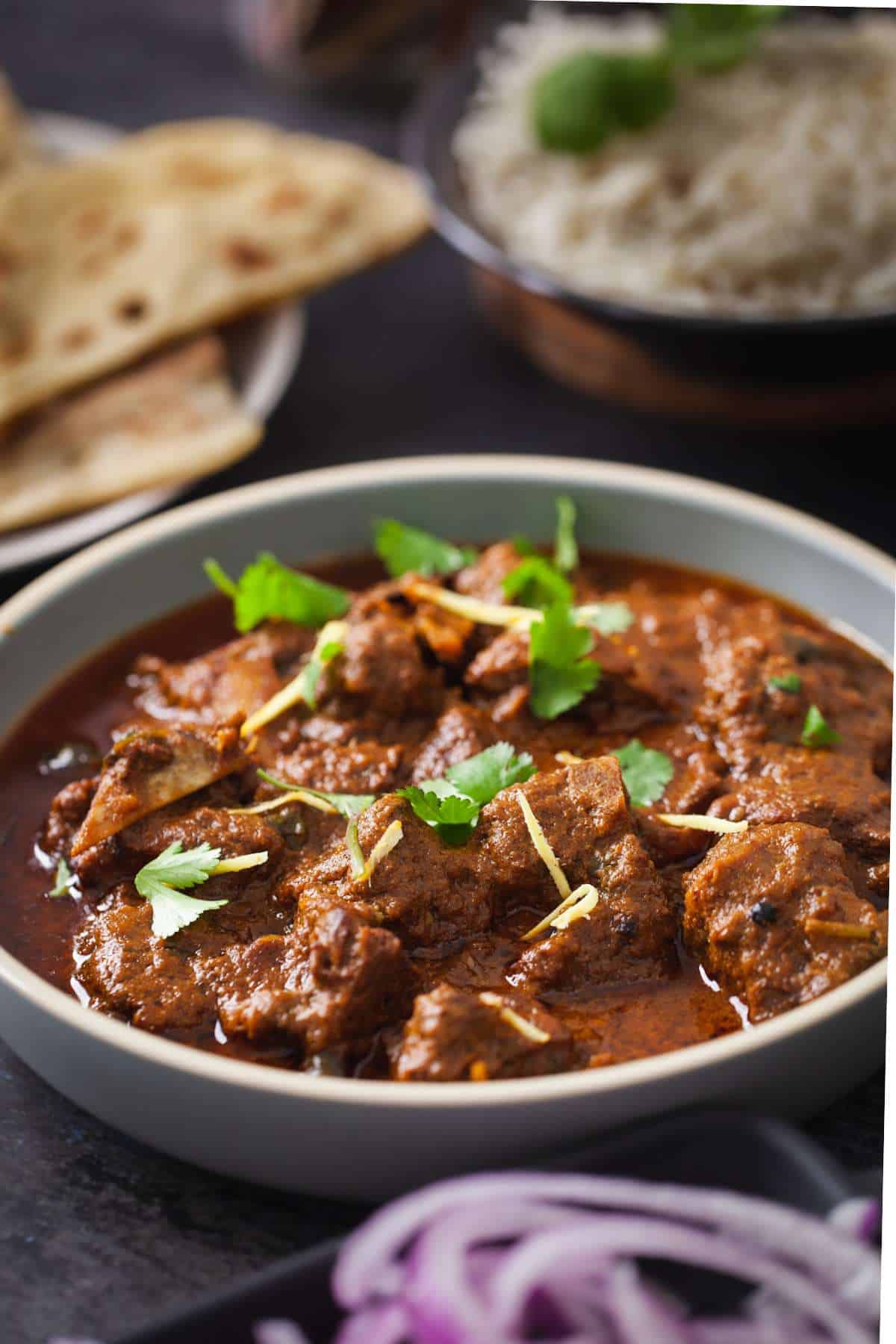 Side view of indian goat curry with rice and flatbread in background. 