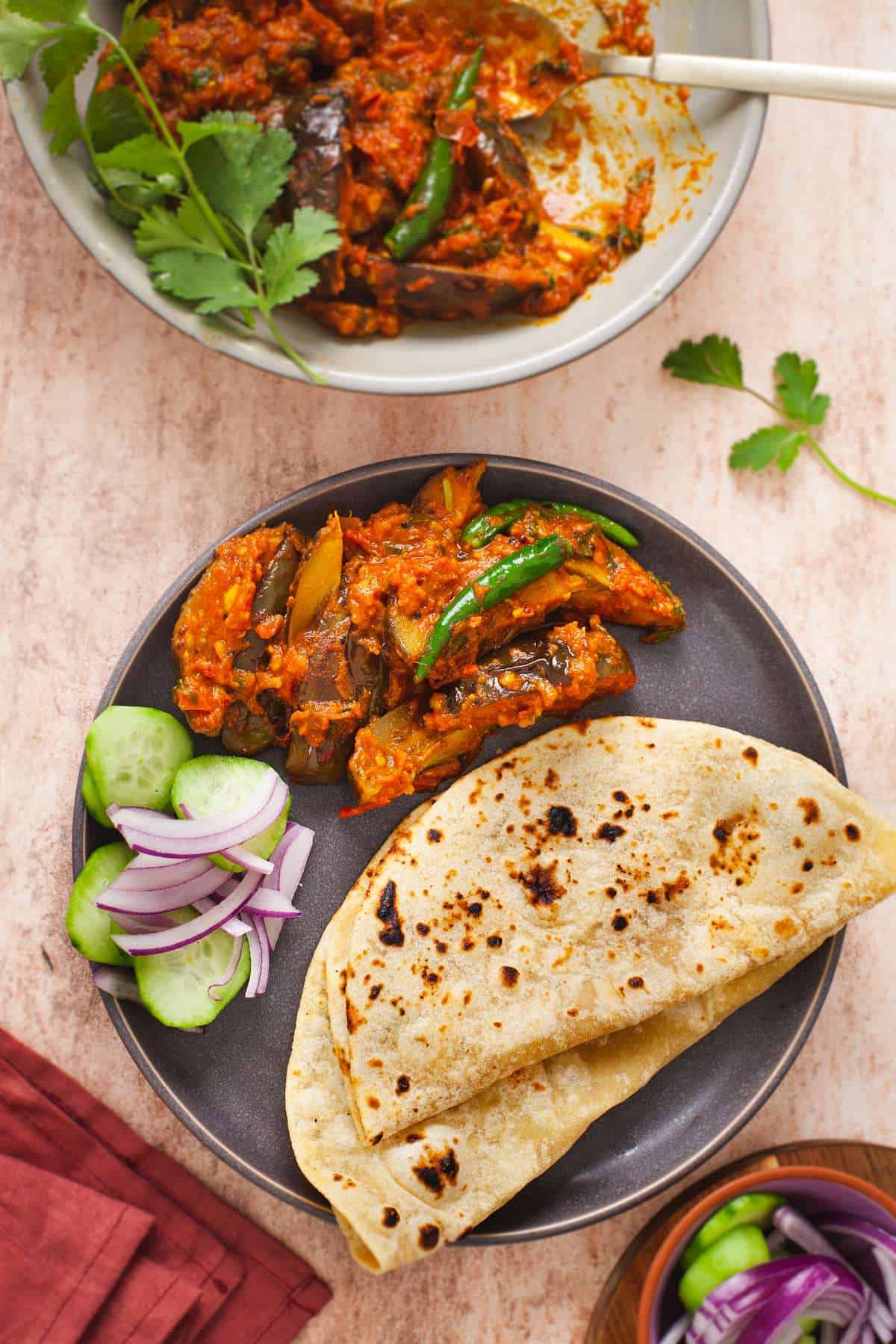 Achari Baingan served with indian flatbread and a salad on a grey stone plate. 