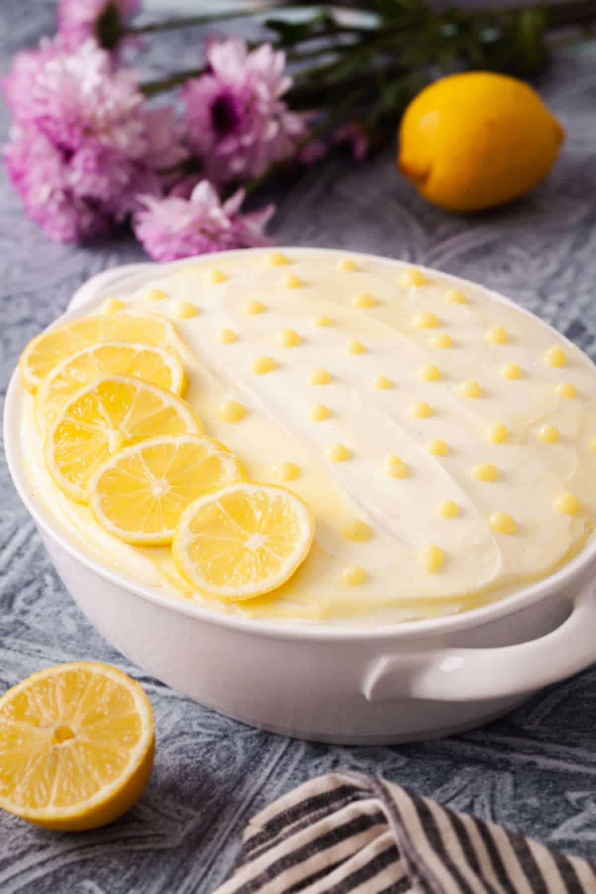 Side shot of lemon tiramisu in an oval ceramic dish with purple flowers in the background.