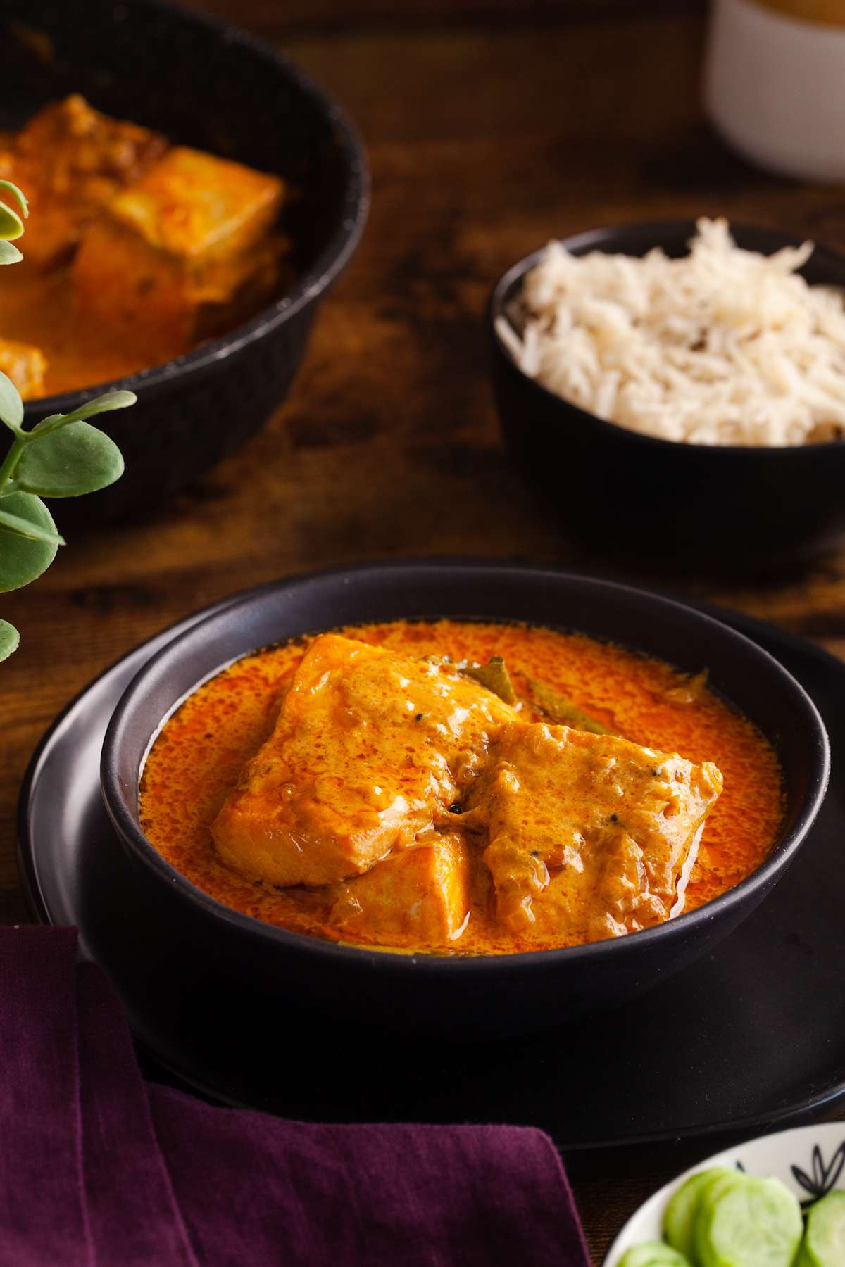 Indian salmon curry in a black bowl placed on a wooden table with white rice in the background. 