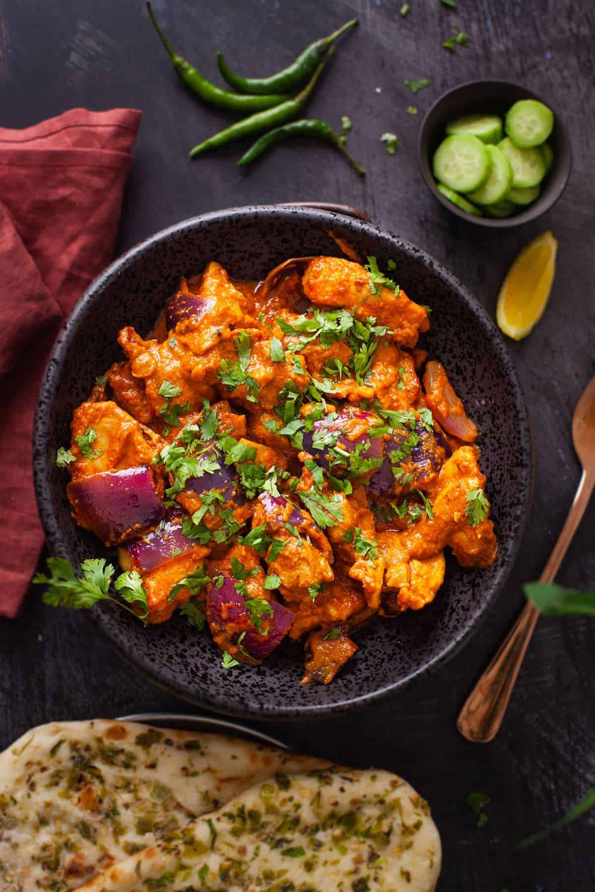 Chicken do pyaza plated in a black colored stone bowl and garnished with cilantro. Served with naan, cut cucumbers and green chillies.