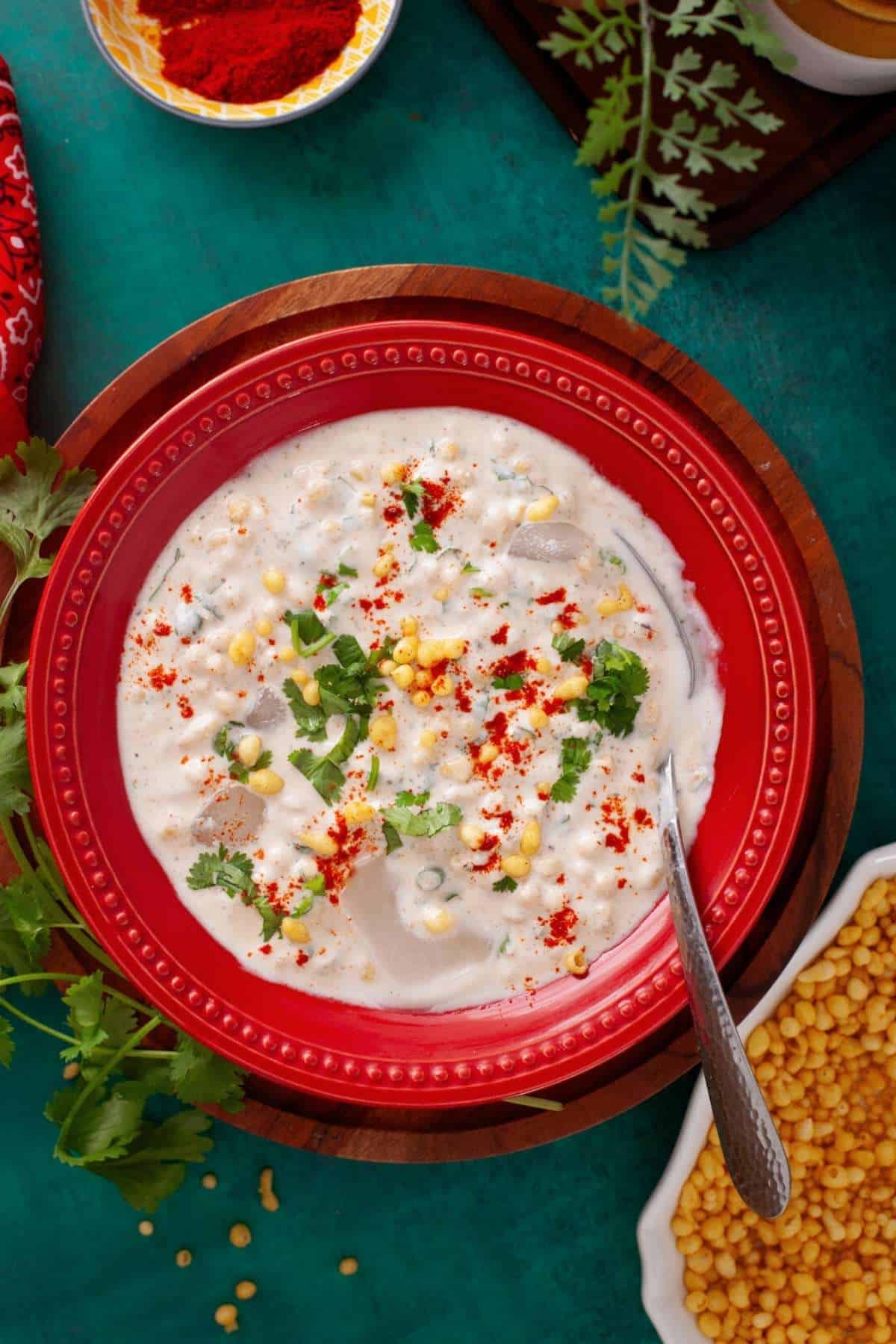 Boondi Raita served in a red dish and garnished with green herbs and sprinkle of red chilli powder.