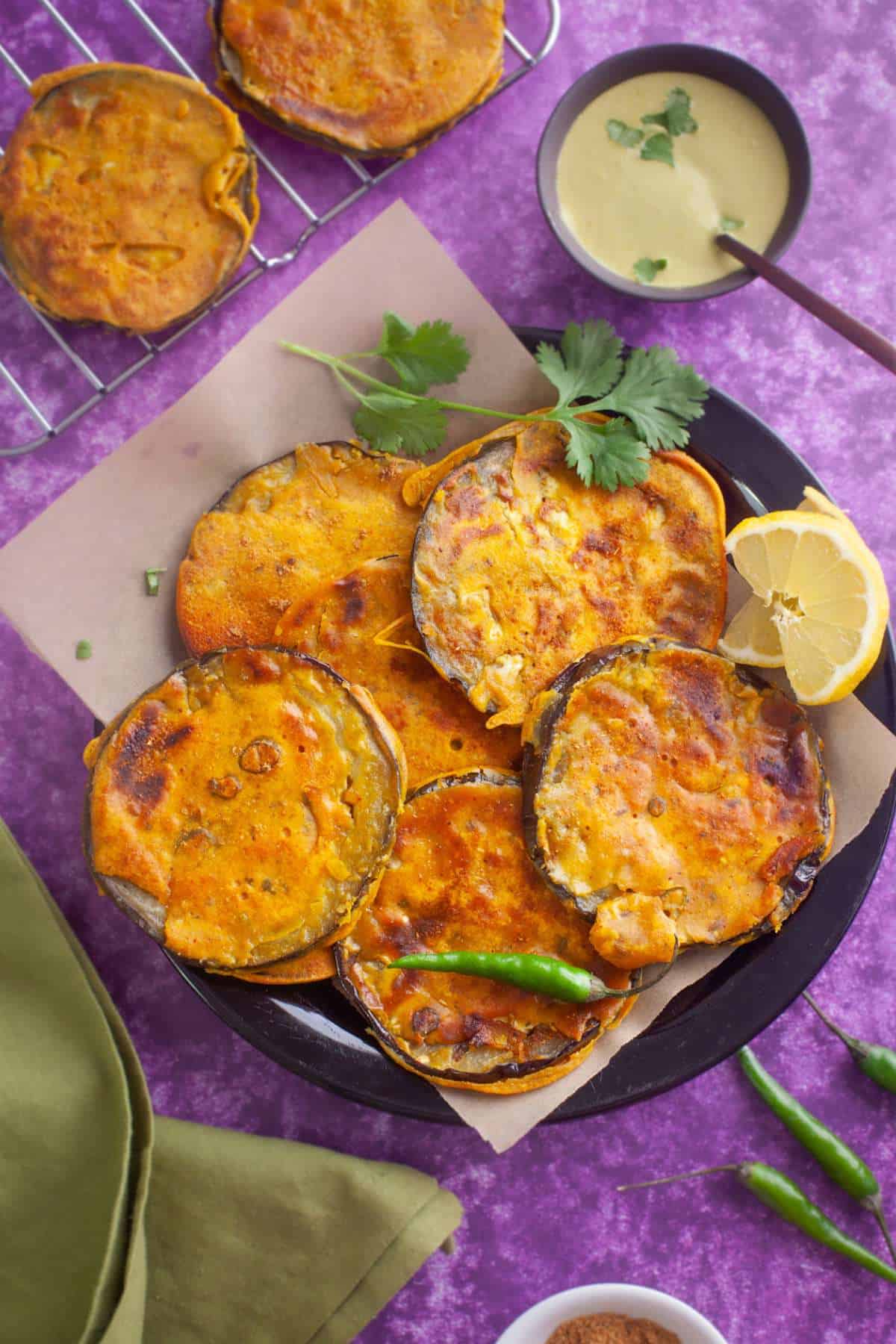 Baingan Pakora served on a black plate with cilantro leaves and a dipping sauce.