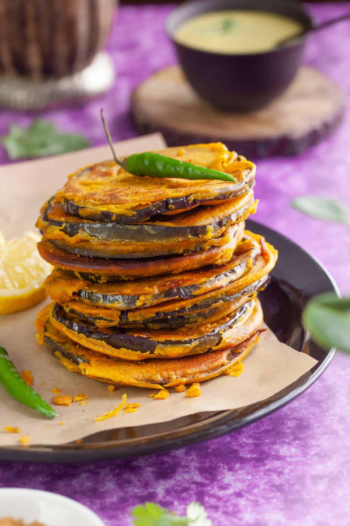 A stack of indian eggplant fritters on a black plate with green chillies and chutney in the background. 