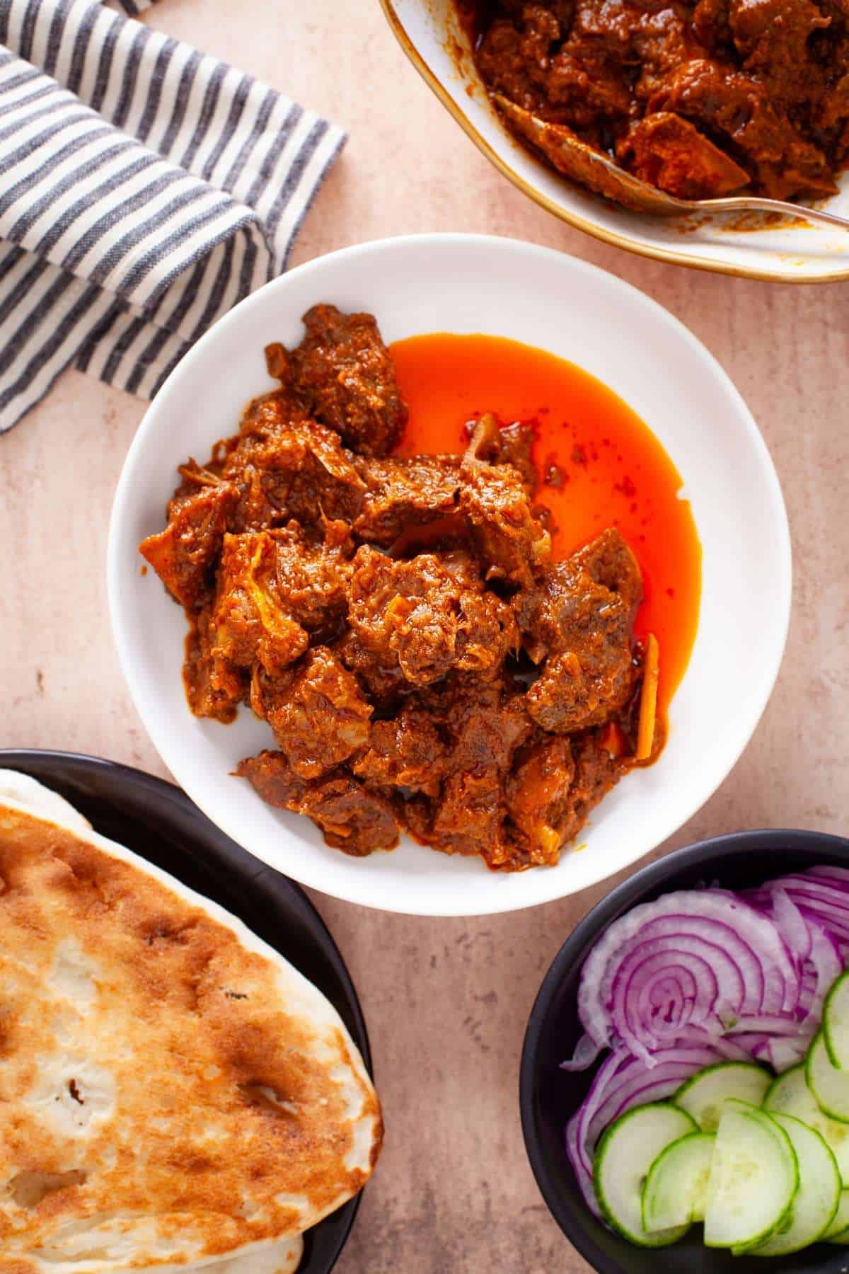 Bhuna gosht in a white bowl with naan, onions and cucumbers.