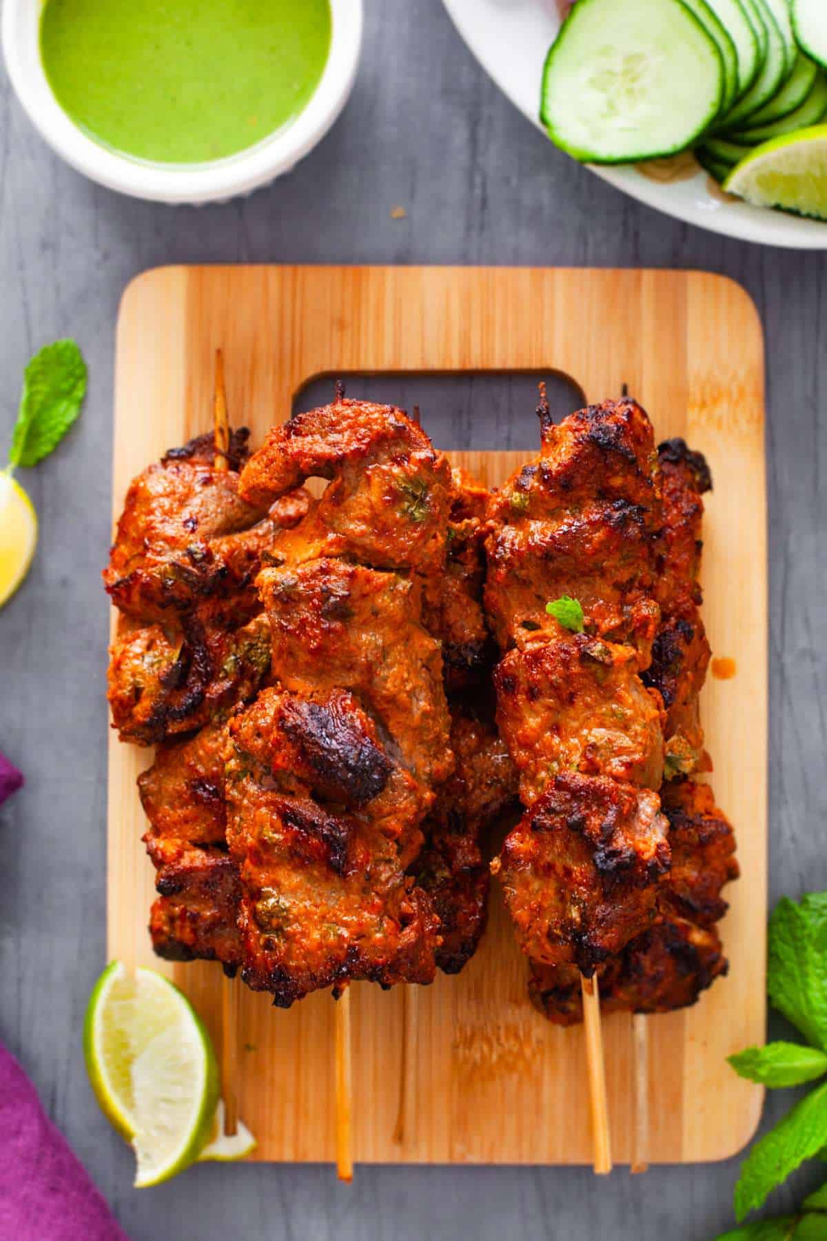 Tandoori Lamb Skewers served on a bamboo board with green chutney and lemon wedges. 