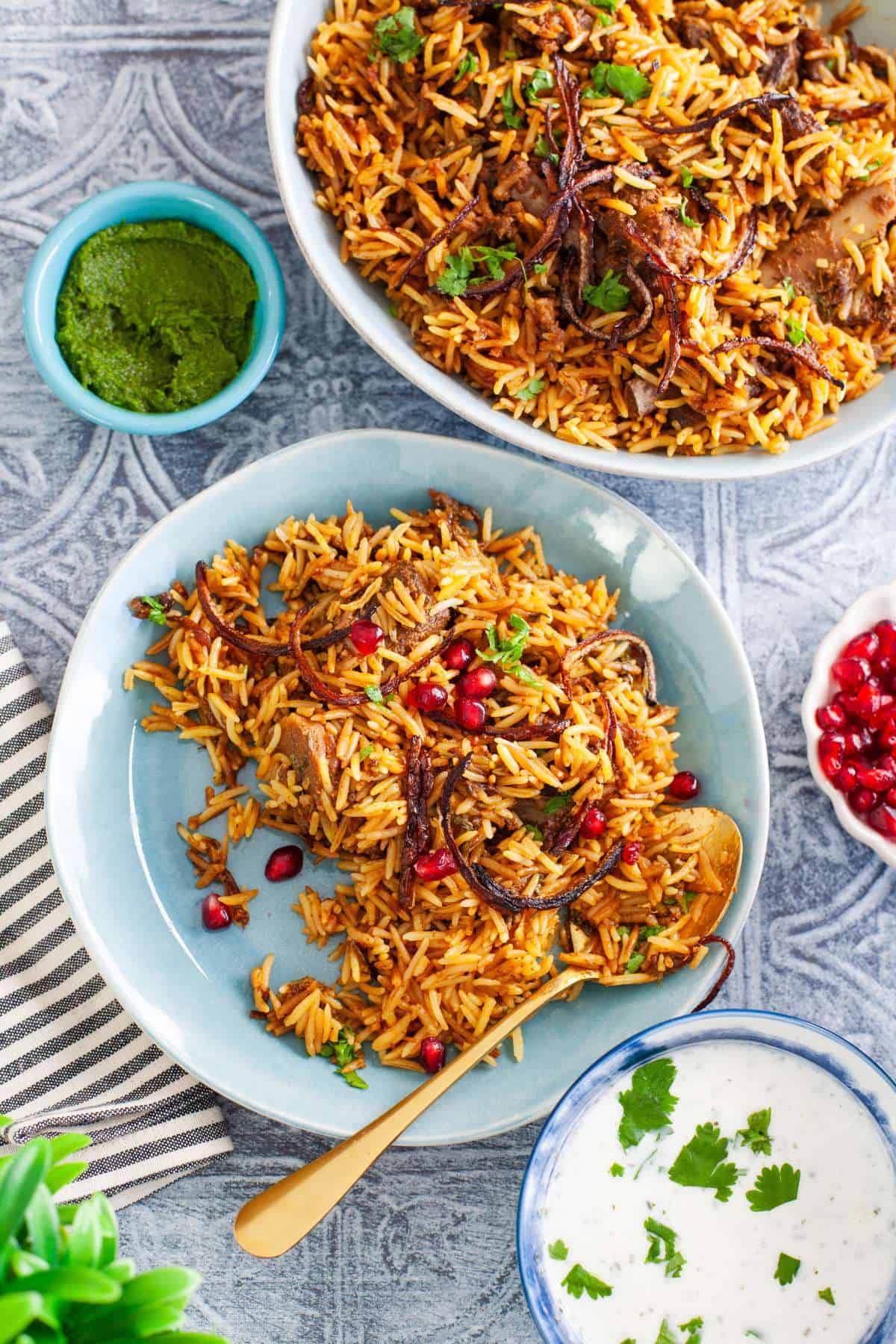 Goat biryani served in a blue plate alongside green chutney and raita.