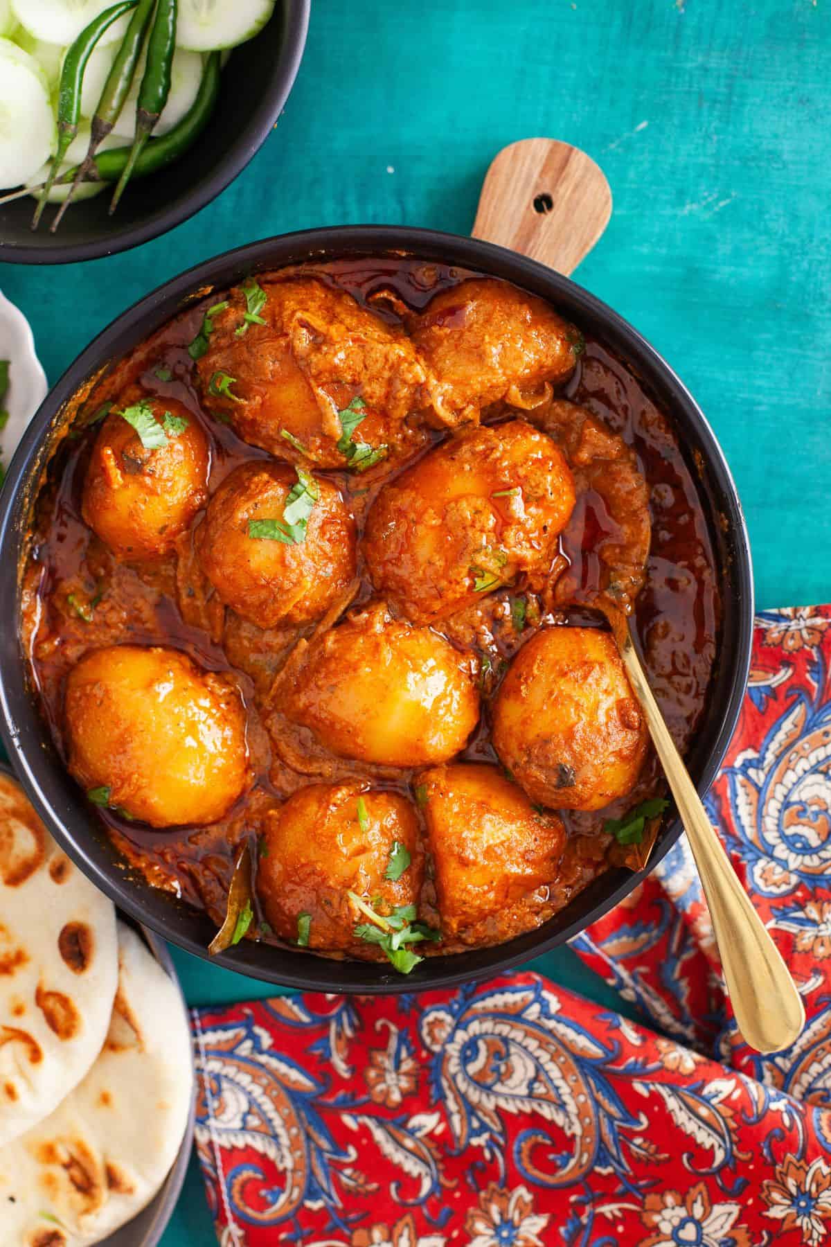 Overhead image of slow cooked potato curry served with cucumber slices and naan.