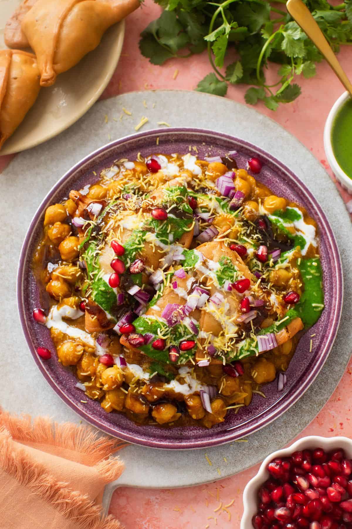Samosa Chaat plated in a purple plate with green chutney, fried samosa and pomegranate on the side.