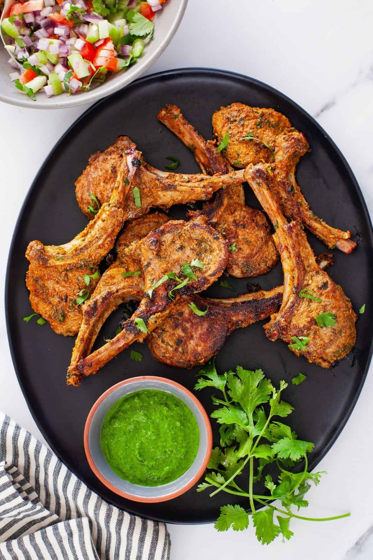 Tandoori Lamb Chops placed in a black serving platter with green chutney and fresh salad. 