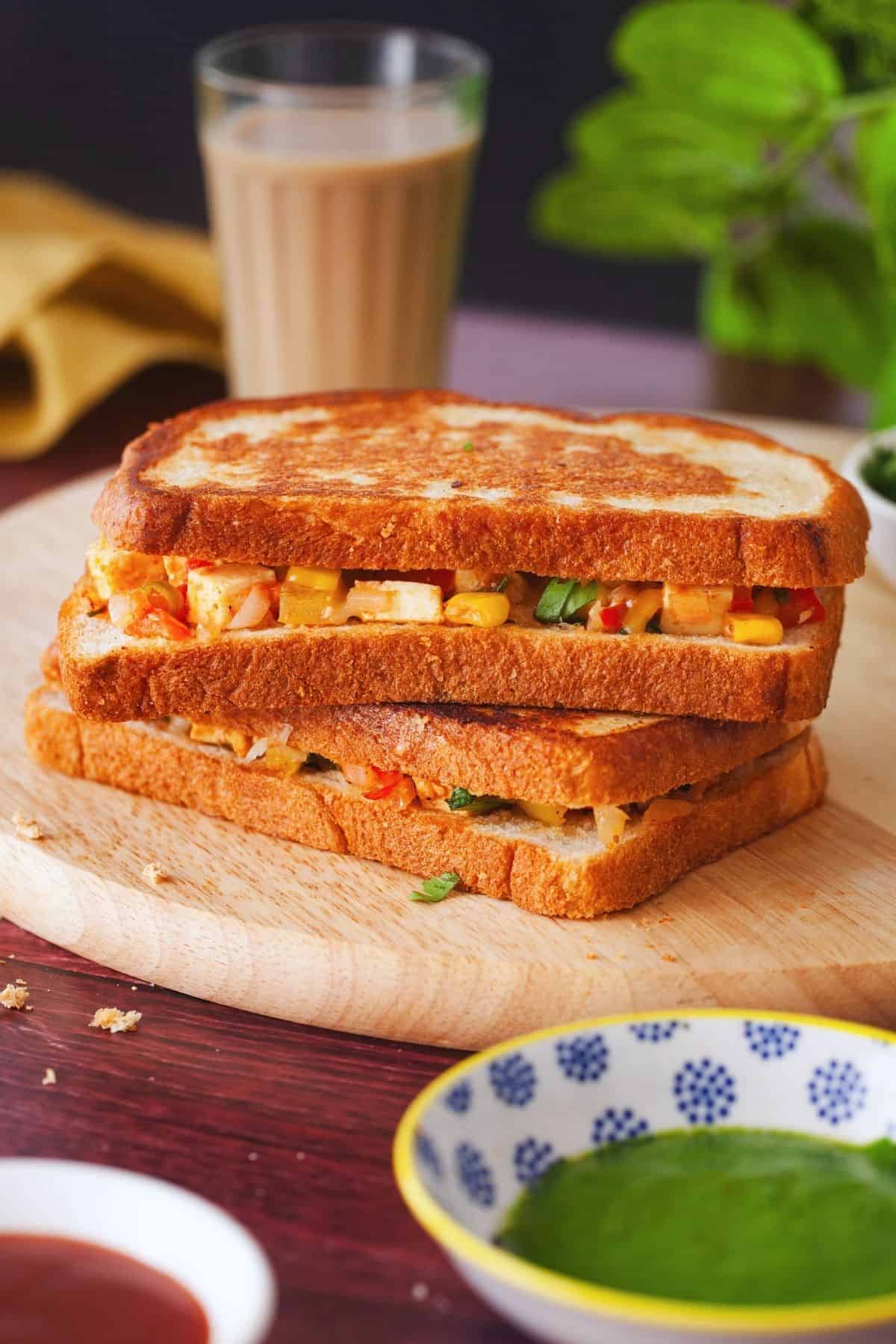 Paneer Sandwich place in a wooden board and served with green chutney and ketchup.