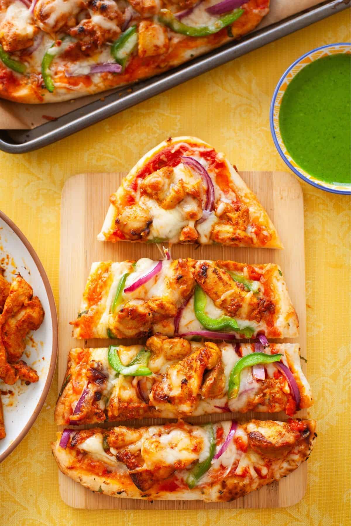Chicken Tikka Pizza sliced and placed on a bambo board with green chutney in side.