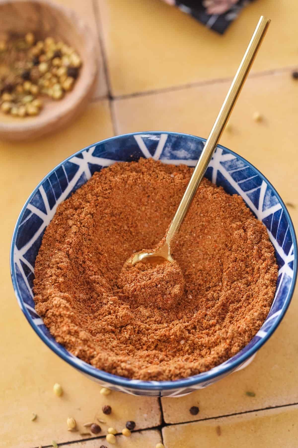 Top view of homemade chaat Masala powder in a blue bowl place on a tiled yellow background.