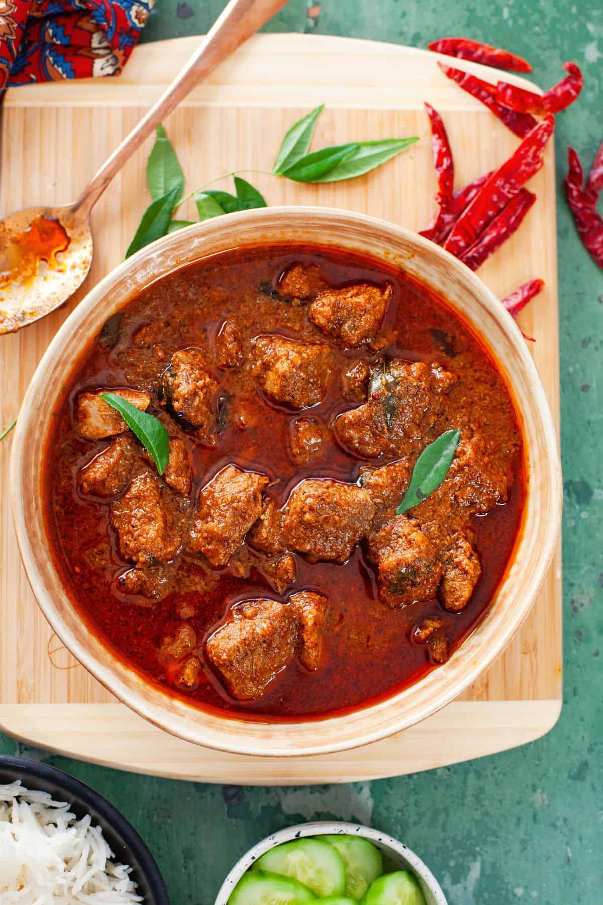Lamb vindaloo plated in a beige ceramic bowl and served with basmati rice and cucumbers.