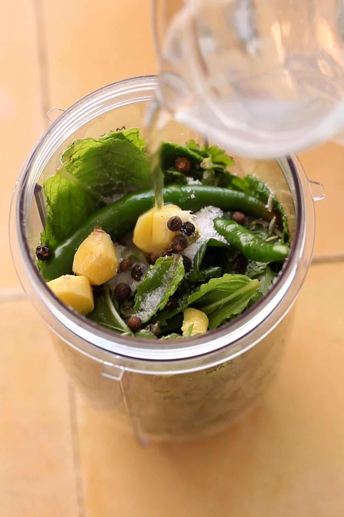 Pouring water over green herbs and spices in a blender jar.