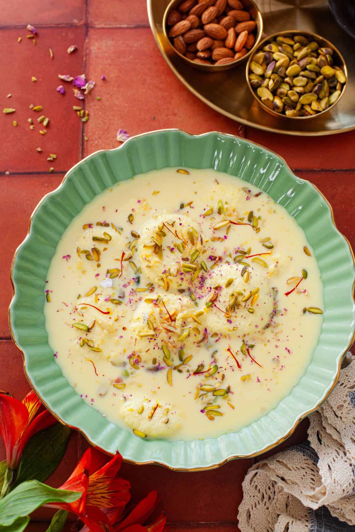 Rasmalai served on a green gold rimmed shallow bowl and garnished with nuts & saffron.