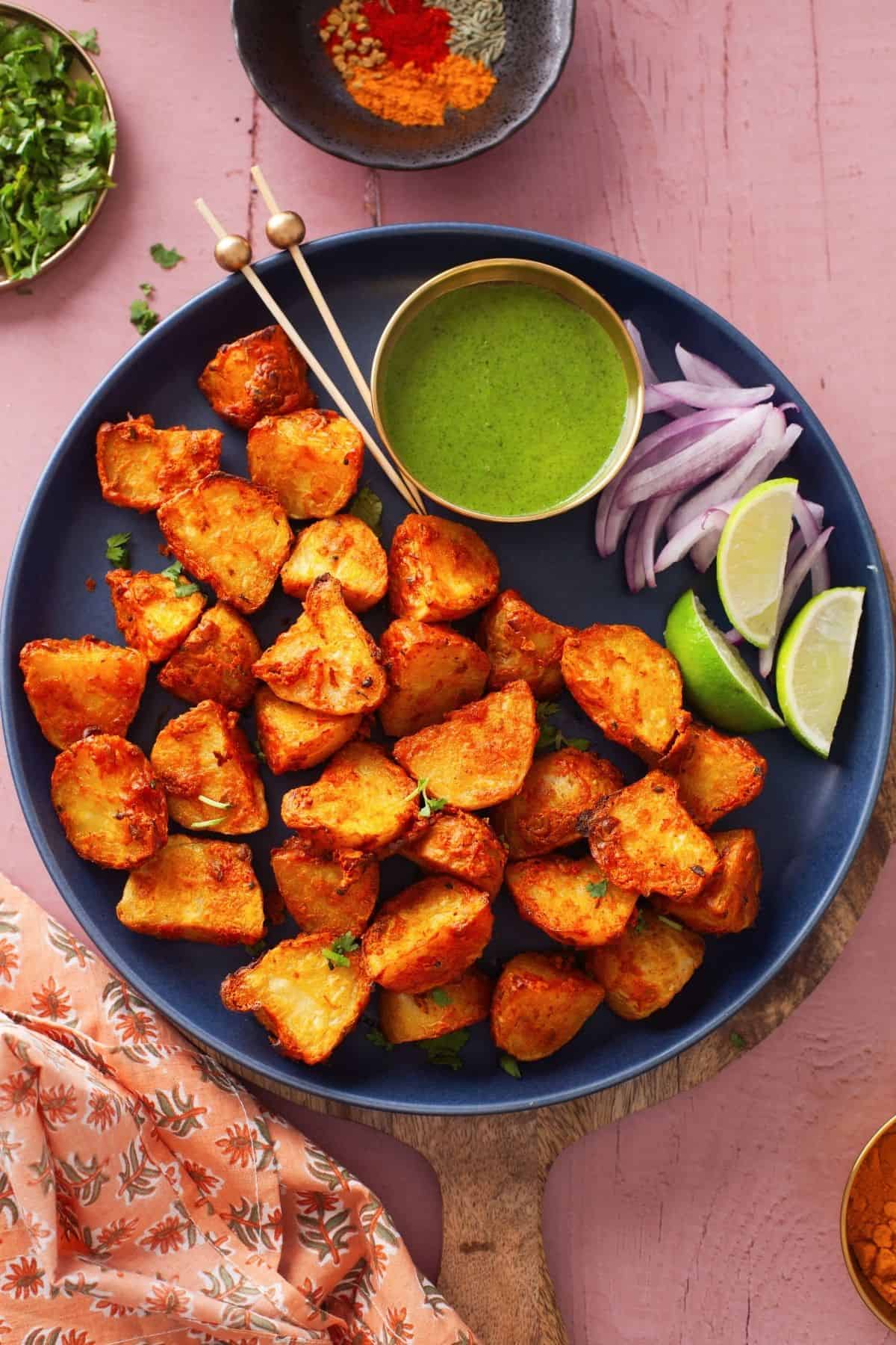 Achari Aloo Tikka Served in a navy blue platter with green chutney, lemon wedges and sliced onion.