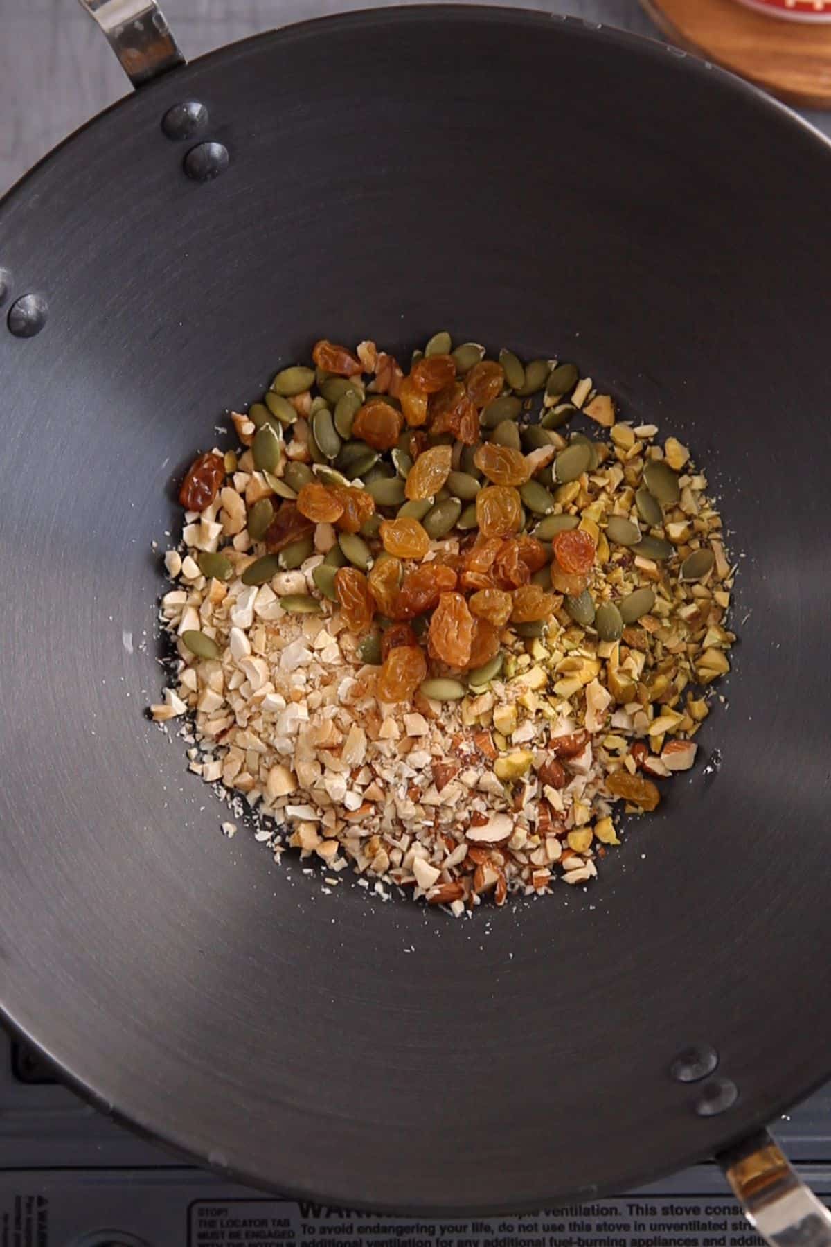 Chopped nuts, raisins, pumpkin seeds in a large black kadai.
