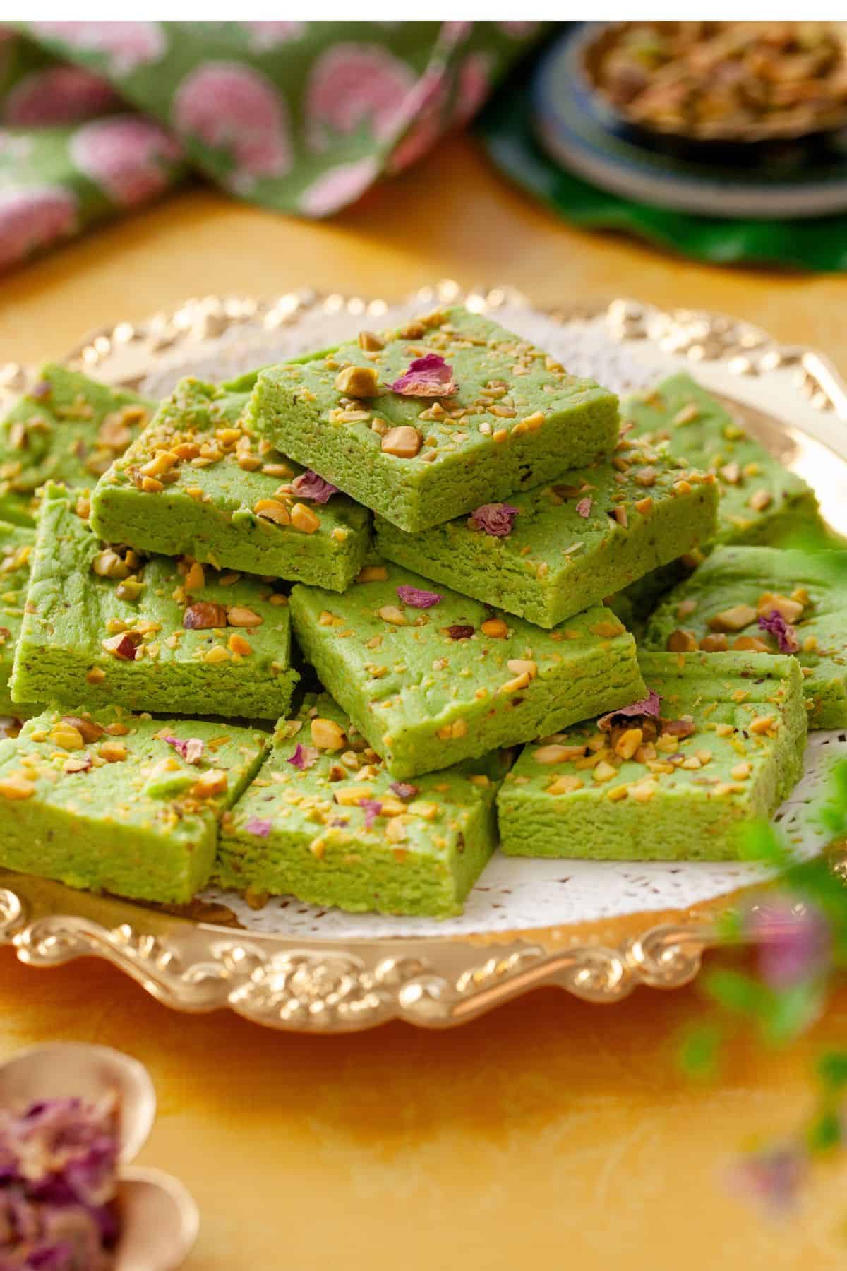 Pistachio burfi squares are placed on a gold plate with pistachios and rose in background.