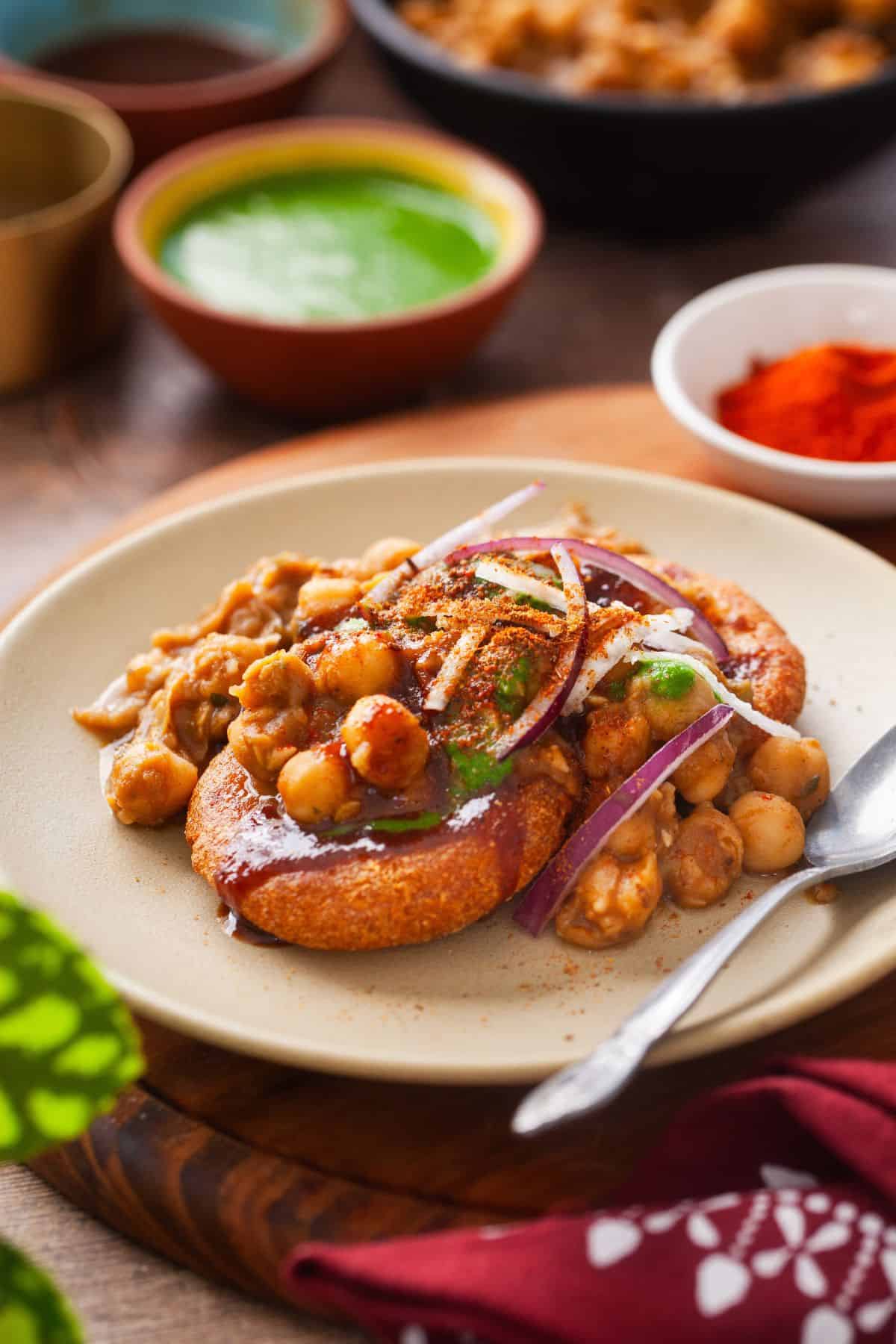 Side image of chole Tikki chaat plated on a mustard colored plate with chutney and spices in background. 