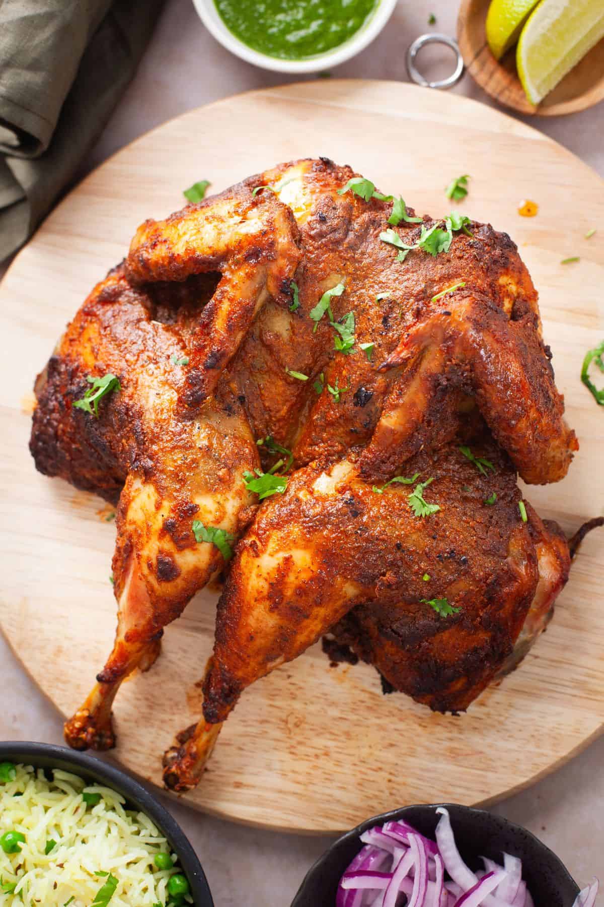 Whole Indian Roast Chicken placed on a wooden board with green chutney, sliced onions and rice pilaf.