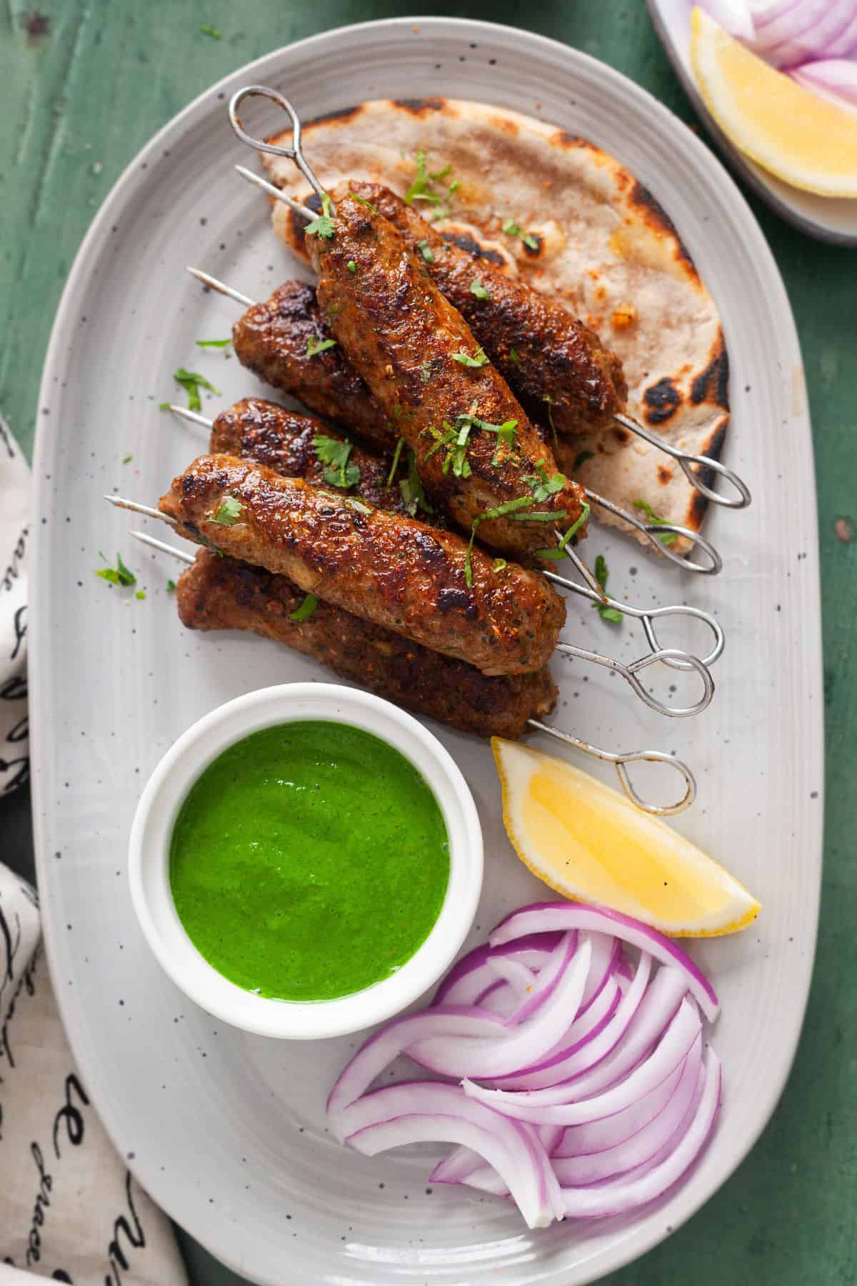 Lamb seekh kebab on metal skewers served on a grew platter with roti, chutney, onion and lemon wedges. 