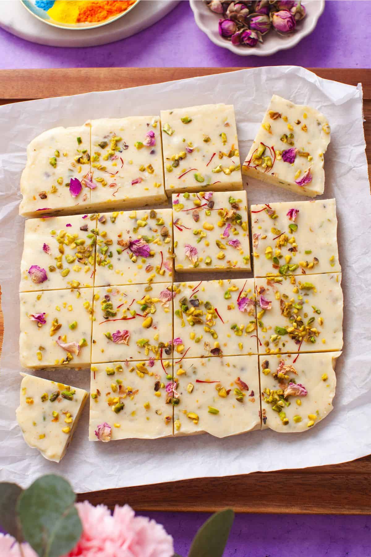 Sliced thandai fudge on parchment paper with plates with colorful holi powders and dried rosebuds are in the background.