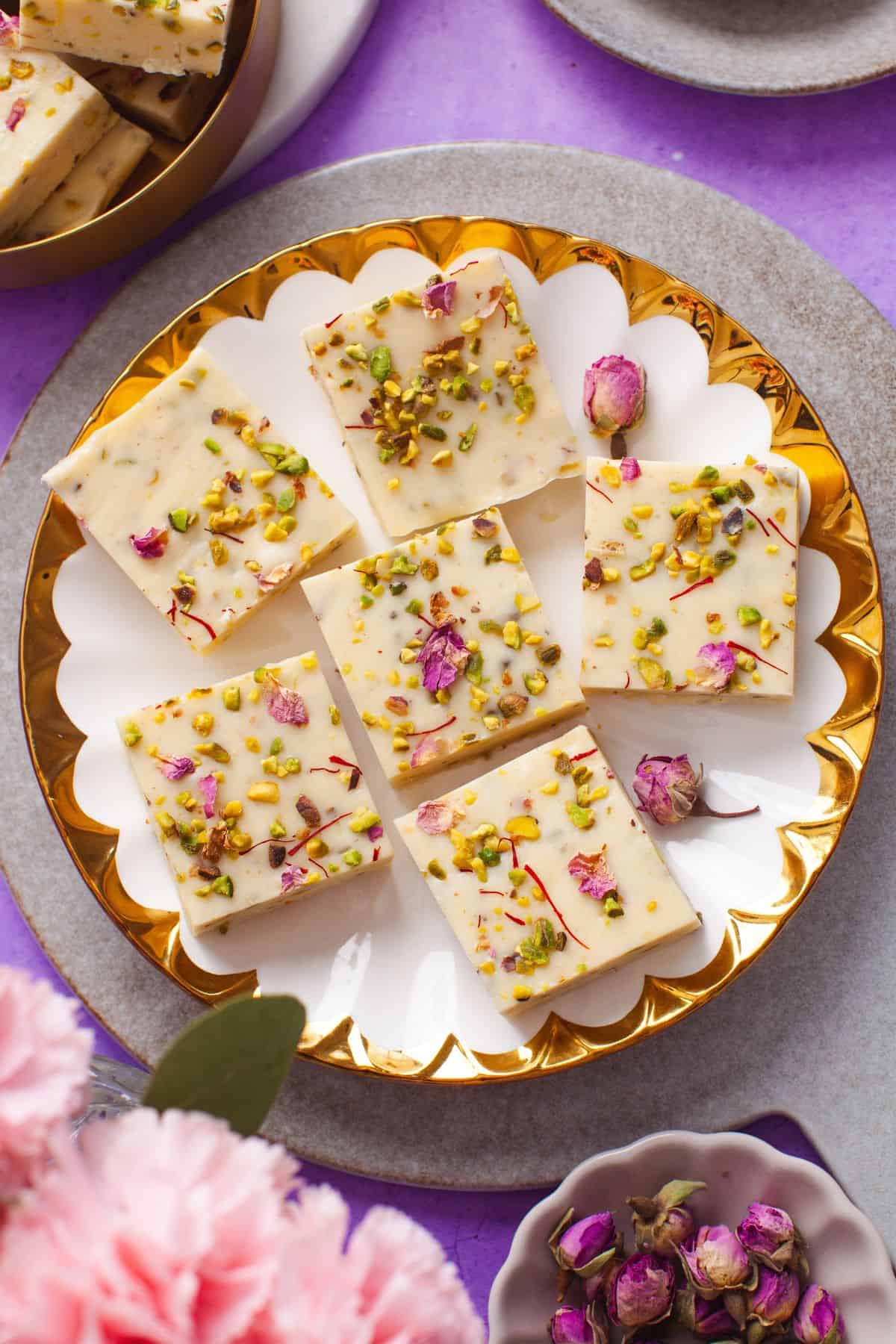 Top view of a white plate with gold trim having six pieces of thandai fudge garnished with chopped pistachios and saffron.