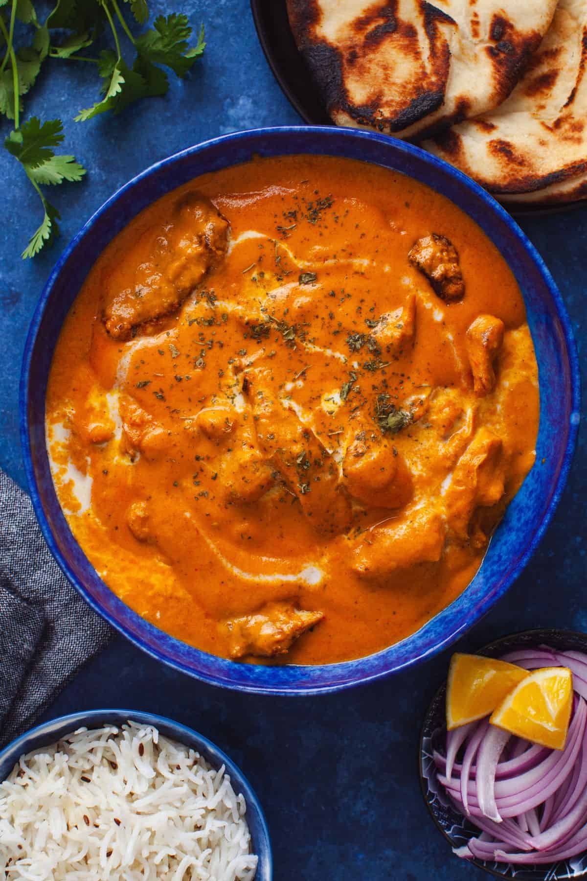 A blue bowl filled with butter chicken curry garnished with dried kasuri methi. Next to it are slices of naan, a bowl of white rice, and a dish with sliced red onions and lemon wedges