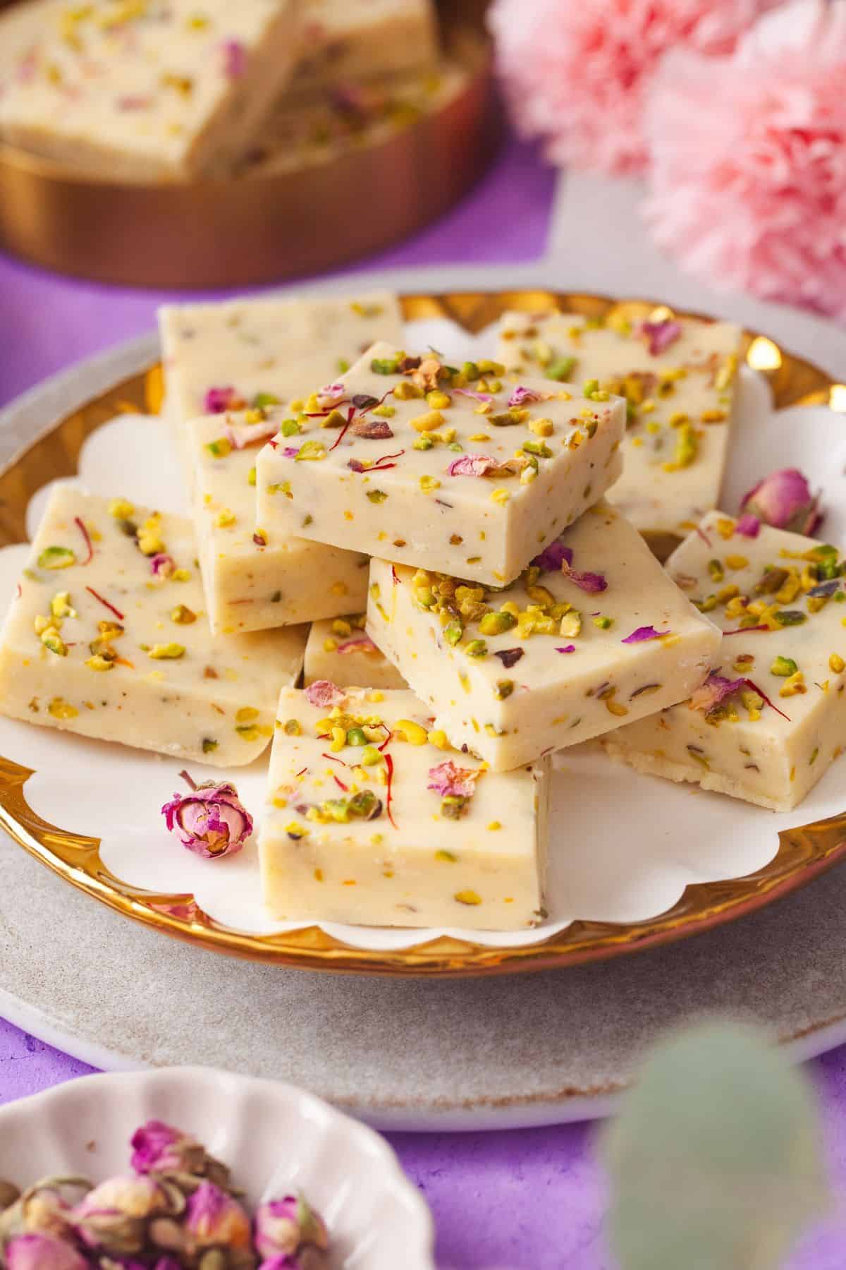 A plate stacked with pieces of creamy white thandai fudge topped with chopped pistachios and rose petals