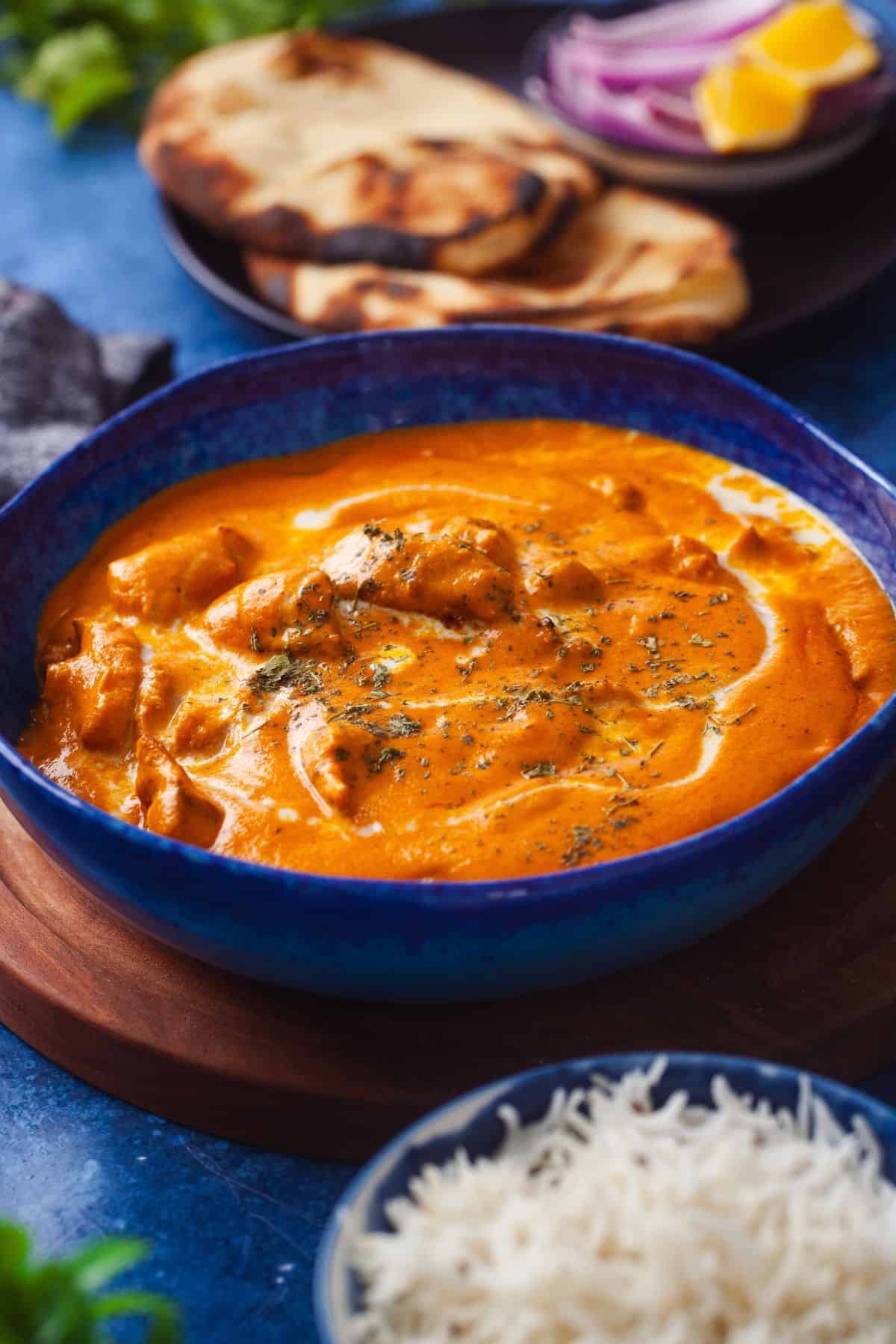 Side image of a blue bowl filled with creamy butter chicken curry sits on a table. Beside it are a plate with sliced naan bread, onions, and lemon wedges.