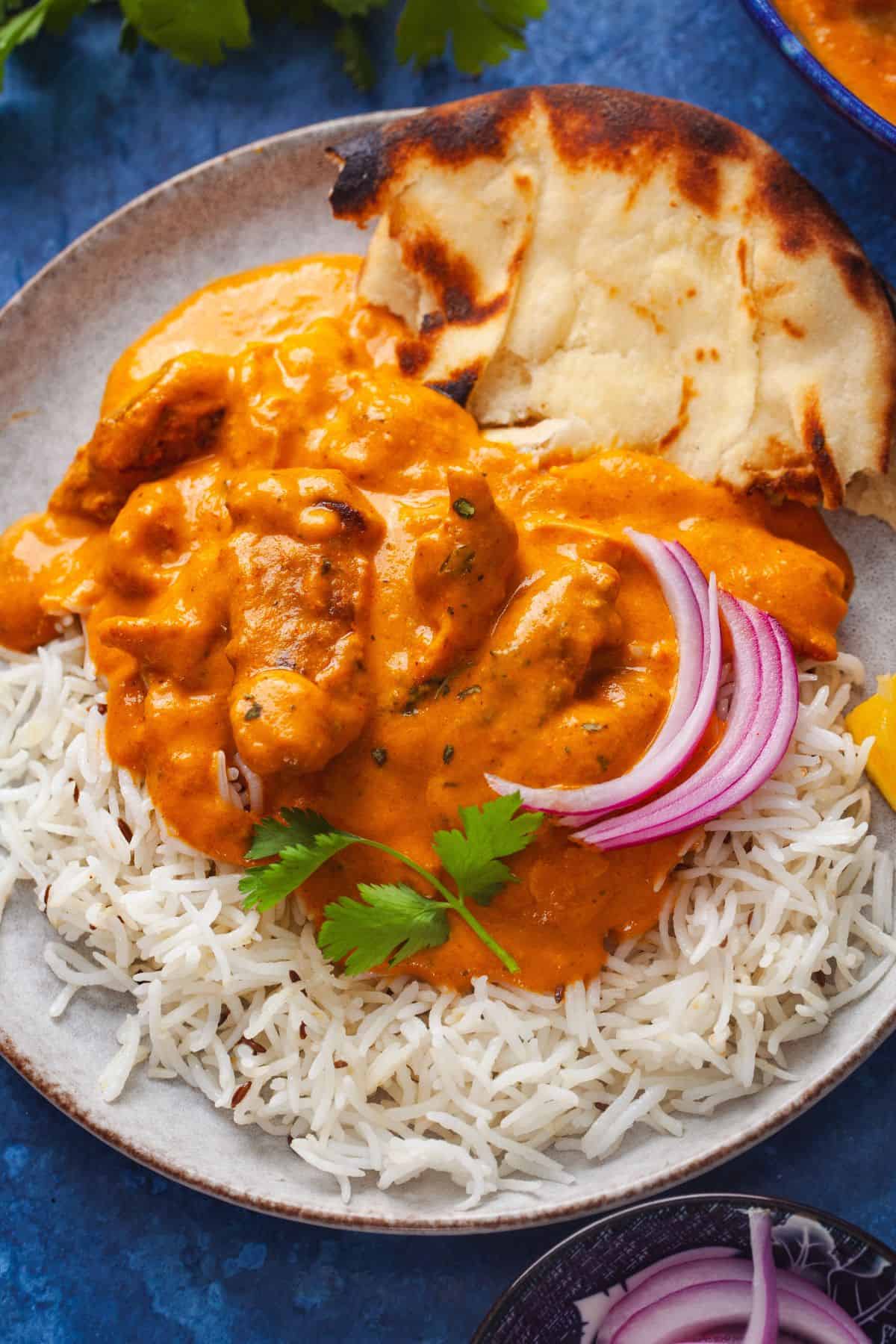 Plate of murgh makhani  with white rice, garnished with cilantro and sliced red onions. Served with a piece of toasted naan bread on a blue background.