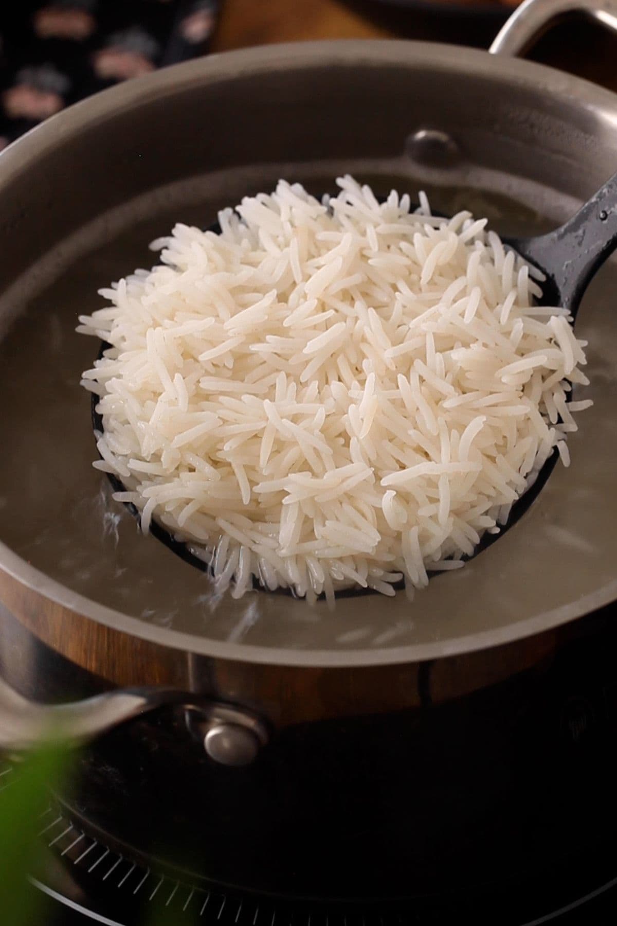 A black slotted spoon holds a serving of cooked white rice above a pot of hot water on a stove.